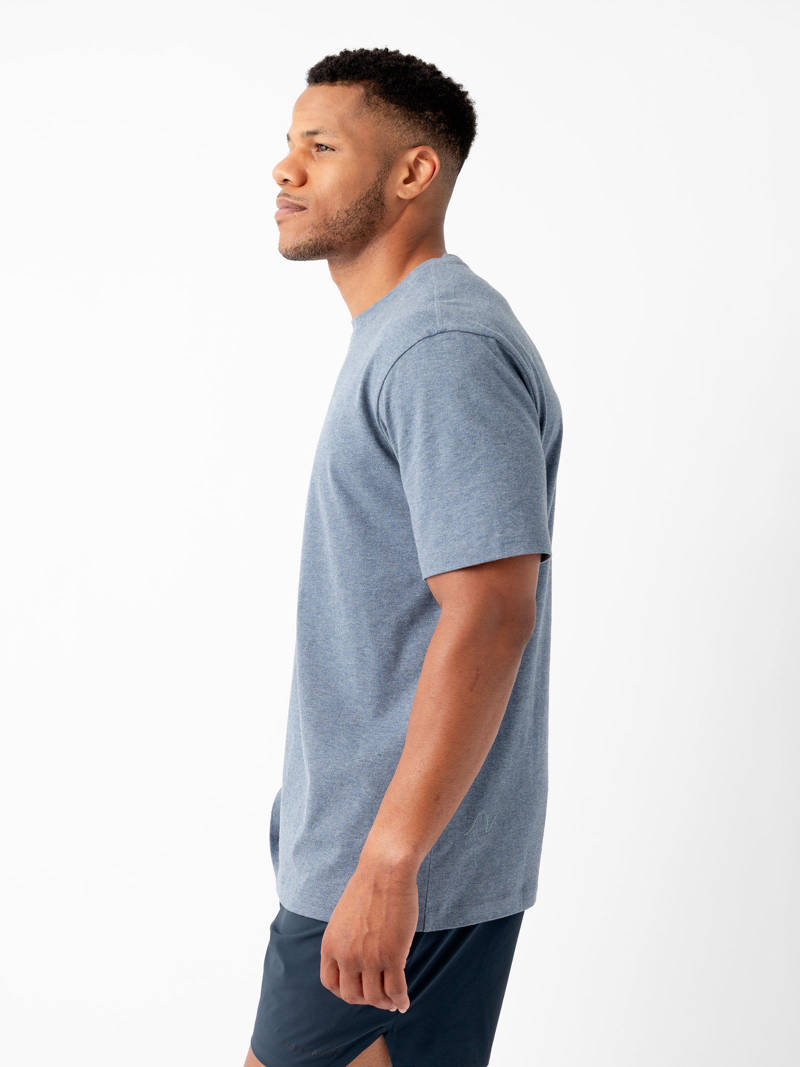 A man stands in profile against a plain white background. He is wearing Cozy Earth's blue Men's All Day Tee and black shorts. His head is turned slightly to the right, and his arms are relaxed at his sides. He has short hair and a neatly trimmed beard. 