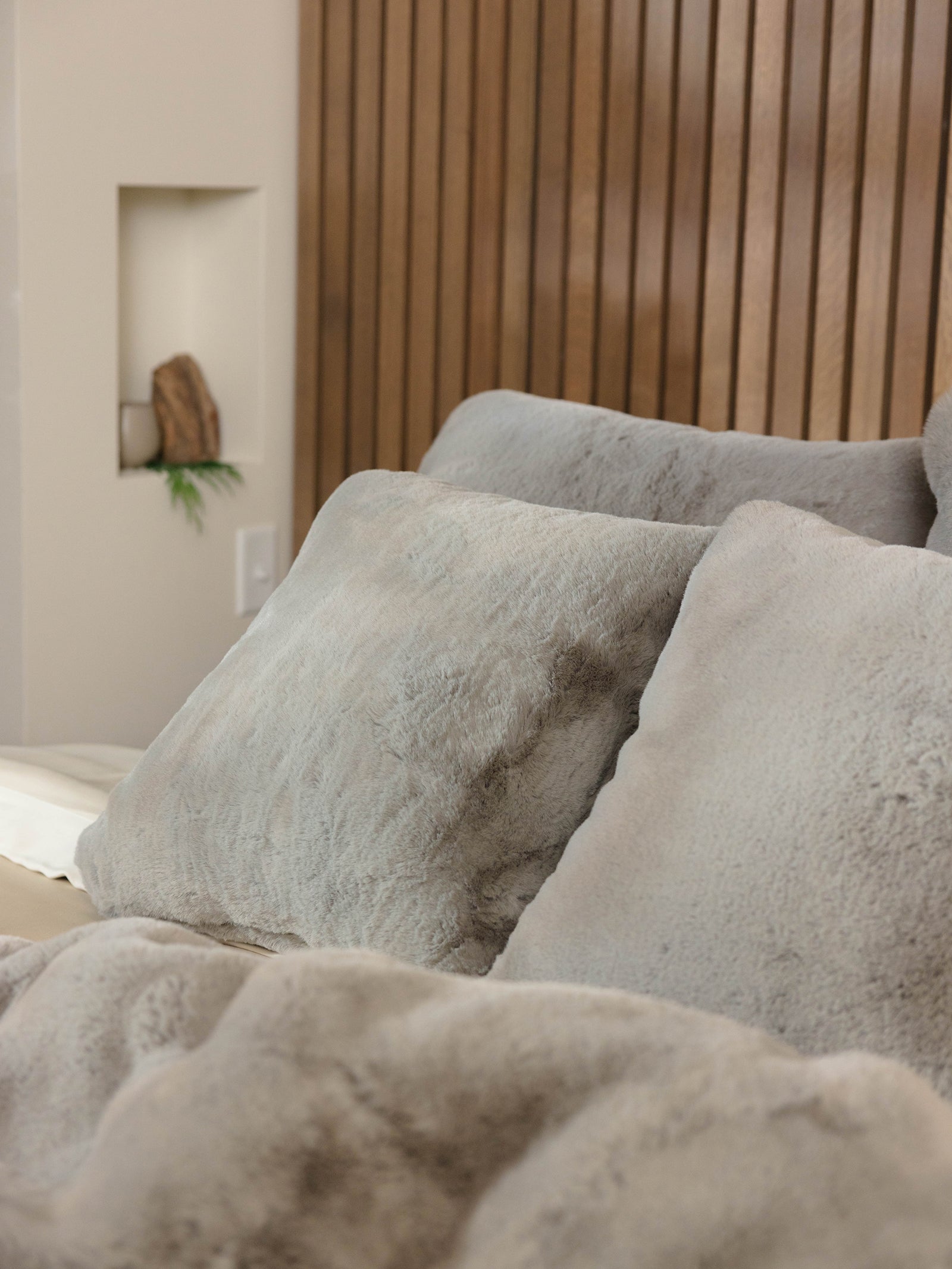 A bed adorned with Cozy Earth's Cuddle Pillow in plush gray, paired with a soft blanket and set against a wooden slatted headboard. A small shelf holding a potted plant can be seen in the background. 