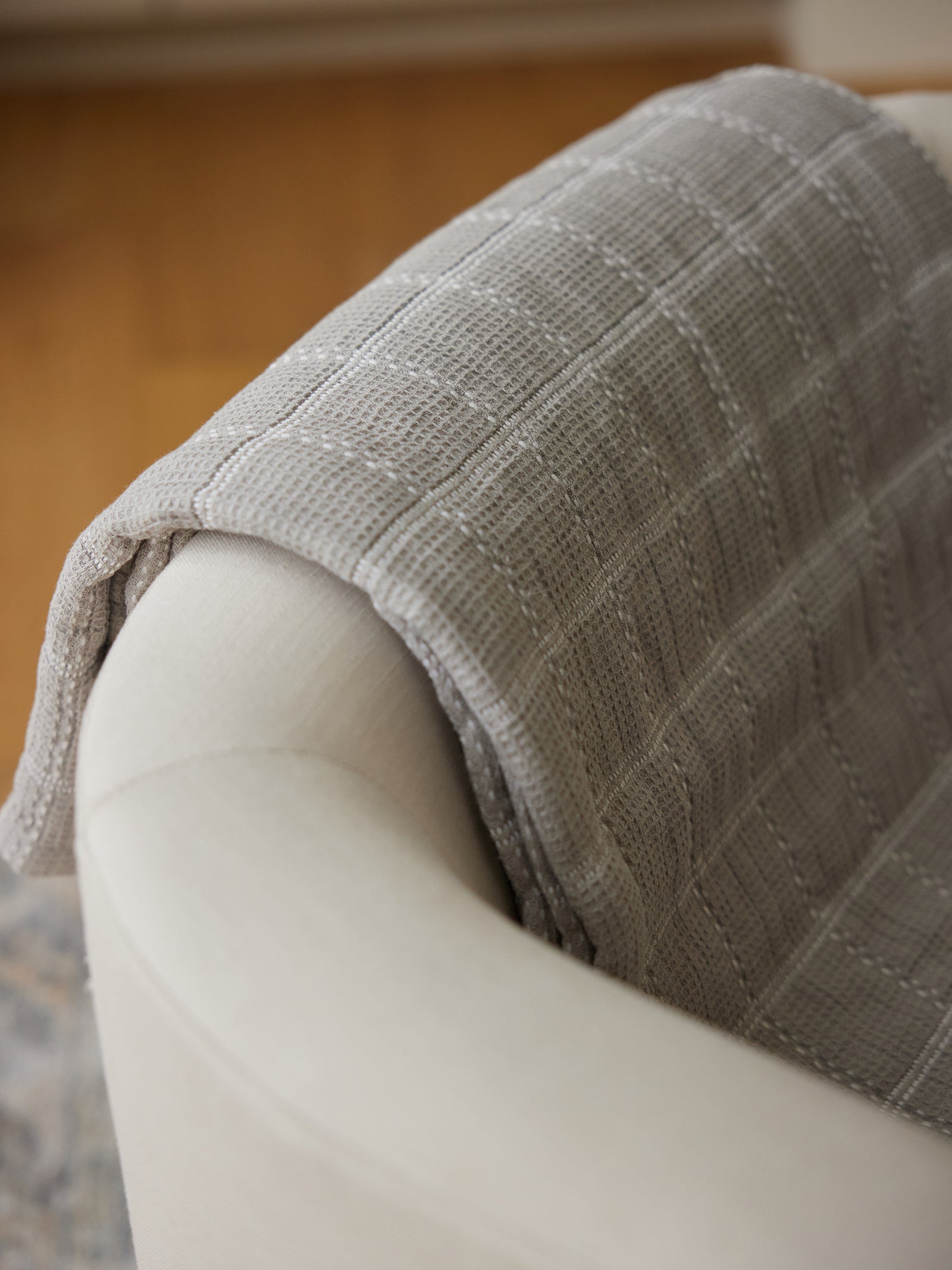 A Waffle Windowpane Blanket from Cozy Earth, in a light gray and textured design, is casually draped over the back of a beige upholstered chair. The chair is positioned on a wooden floor, creating a cozy and inviting atmosphere. 