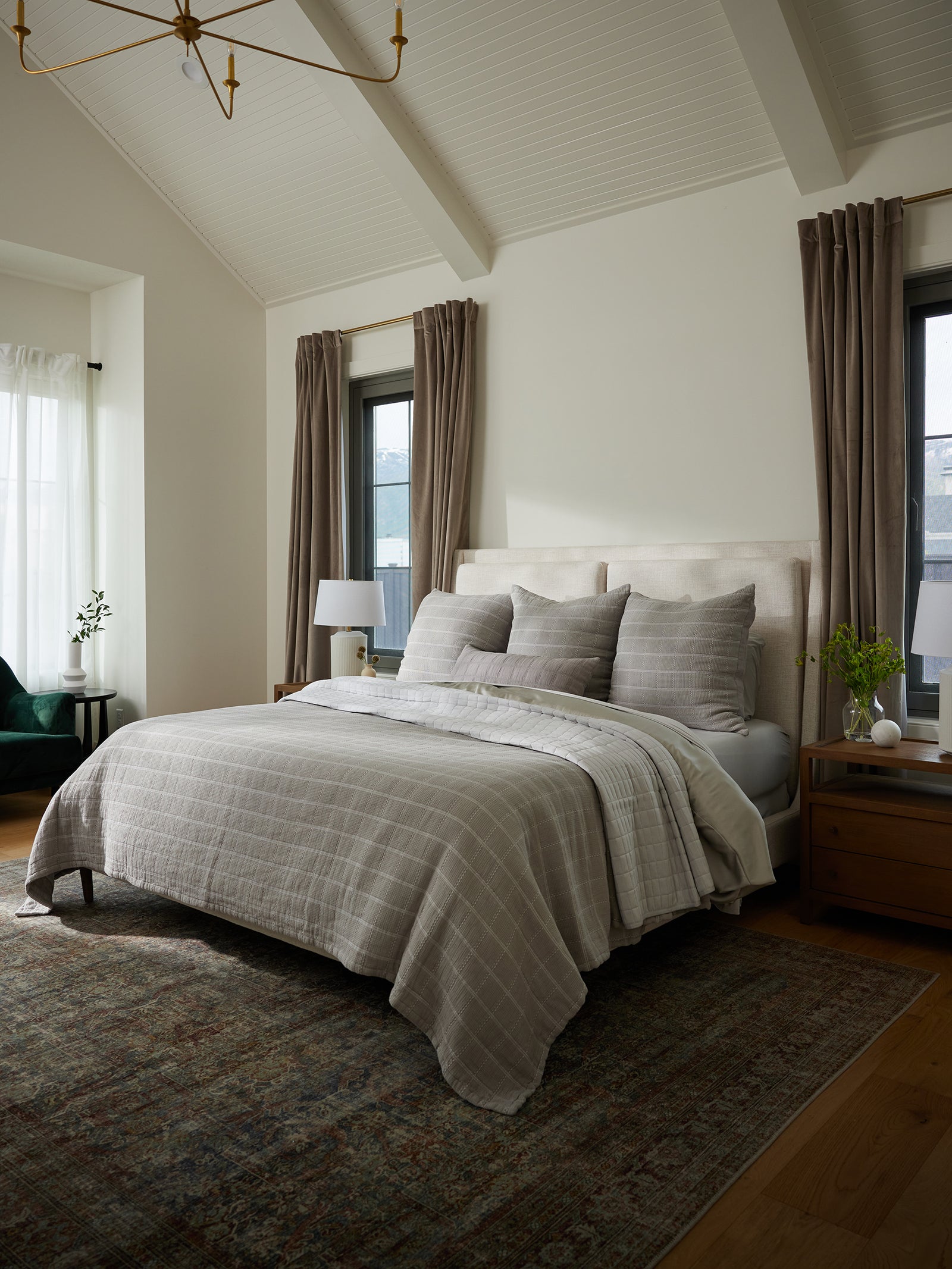 A cozy bedroom showcases a neatly made bed with gray bedding and multiple Waffle Windowpane Pillows by Cozy Earth. The bed is flanked by two wooden nightstands, each topped with matching white lamps. The room includes two tall, narrow windows dressed with long beige curtains, a patterned rug, and a high vaulted white ceiling. 