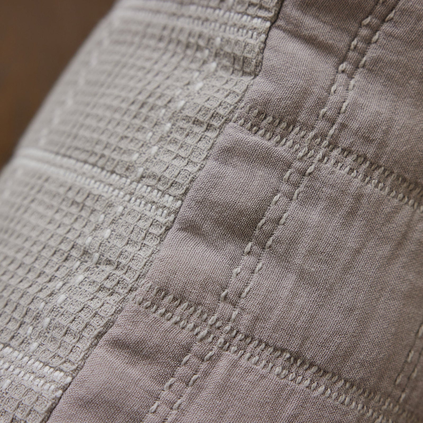 Close-up of the Waffle Windowpane Pillow by Cozy Earth, showcasing a textile surface with a combination of woven and stitched patterns. The fabric features a grid-like texture with raised lines and squares, intersected by parallel rows of stitching. The material appears to be a light grey color. 