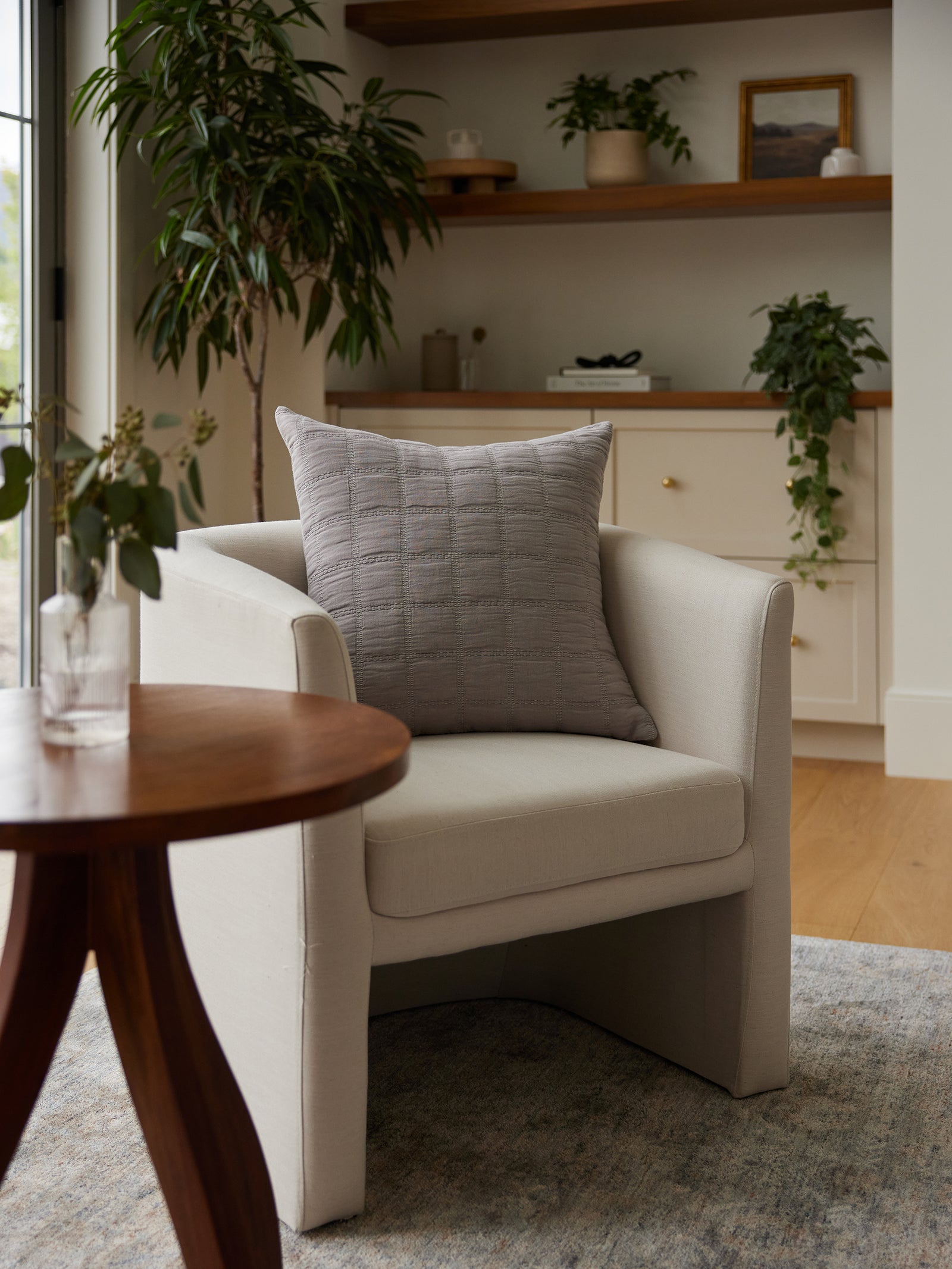 A cozy living room corner featuring a light beige armchair with a Waffle Windowpane Pillow by Cozy Earth, sitting next to a round wooden side table with a vase of fresh flowers. Shelves with plants, decorative items, and a white cabinet are in the background. 