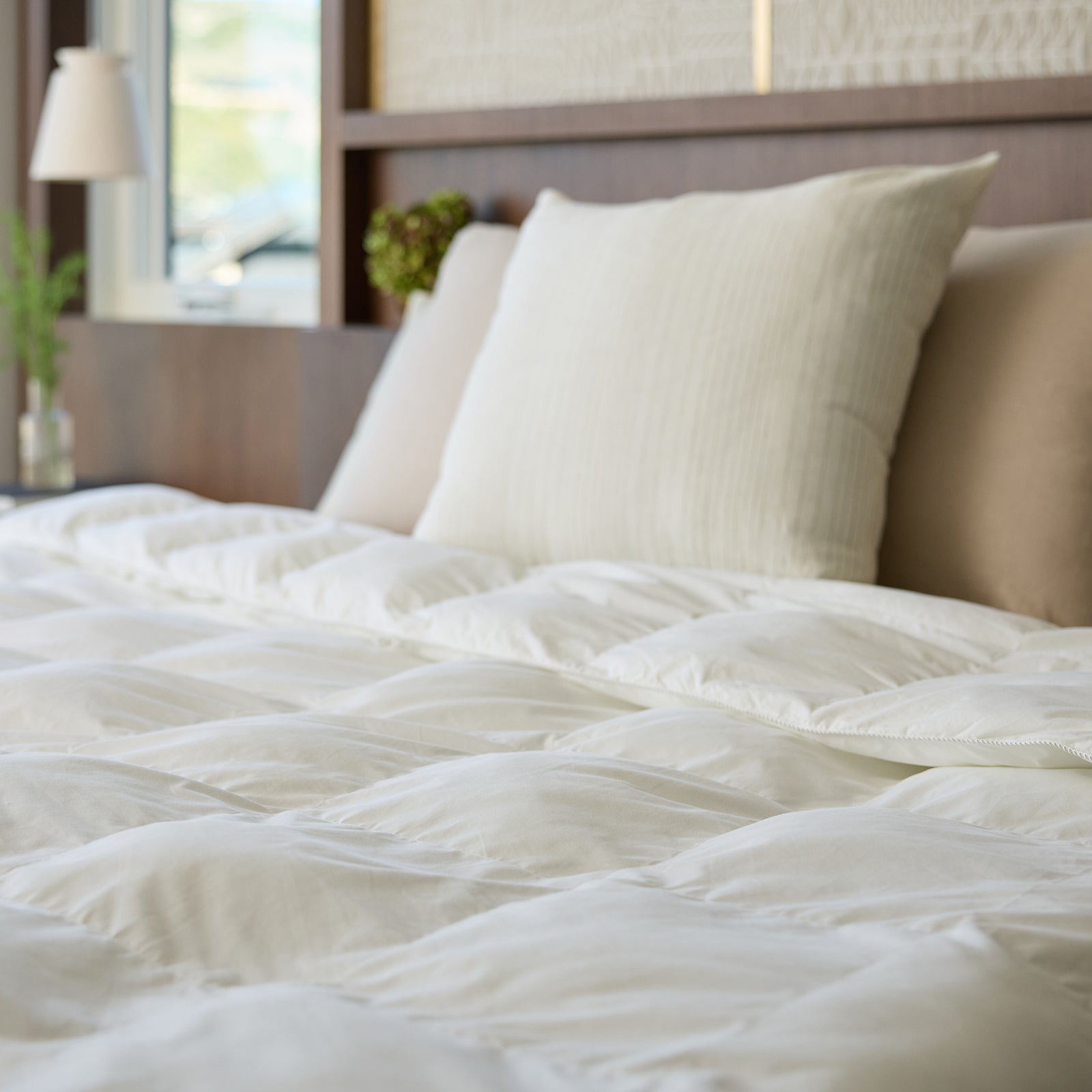 A neatly made bed features the Cozy Earth Down Alternative Comforter in a cozy, sunlit bedroom, with cream-colored pillows adding to the warmth. A small, modern lamp and a green plant rest on the wooden bedside table.