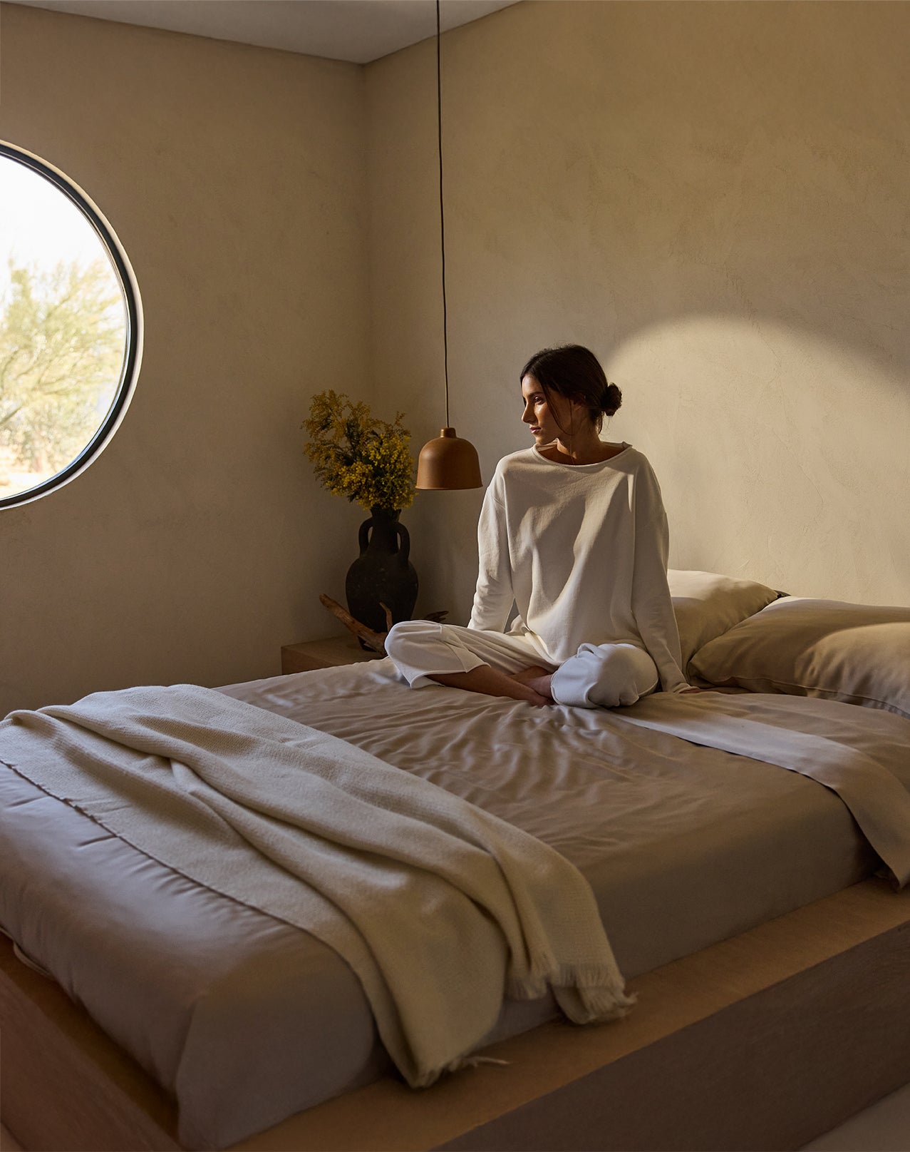 A woman sits cross-legged on a neatly made bed, adorned with a Cozy Earth Bamboo Flat Sheet. She wears white loungewear and gazes out of a round window that floods warm light into the minimalistic, neutral-toned room featuring a round lamp and a vase with flowers. 