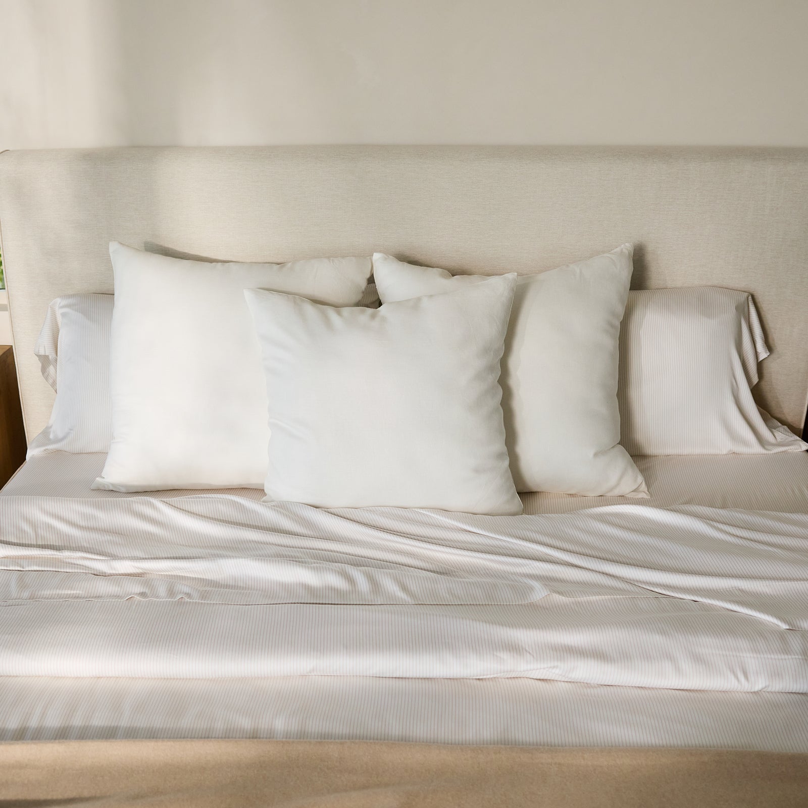 The bed features a neat arrangement of four pillows encased in Bamboo Pillowcases by Cozy Earth, set against an elegant cream-colored headboard. The ensemble is dressed with pristine white sheets and a sleek beige blanket, all warmly illuminated by soft natural light to produce a serene and welcoming atmosphere. 