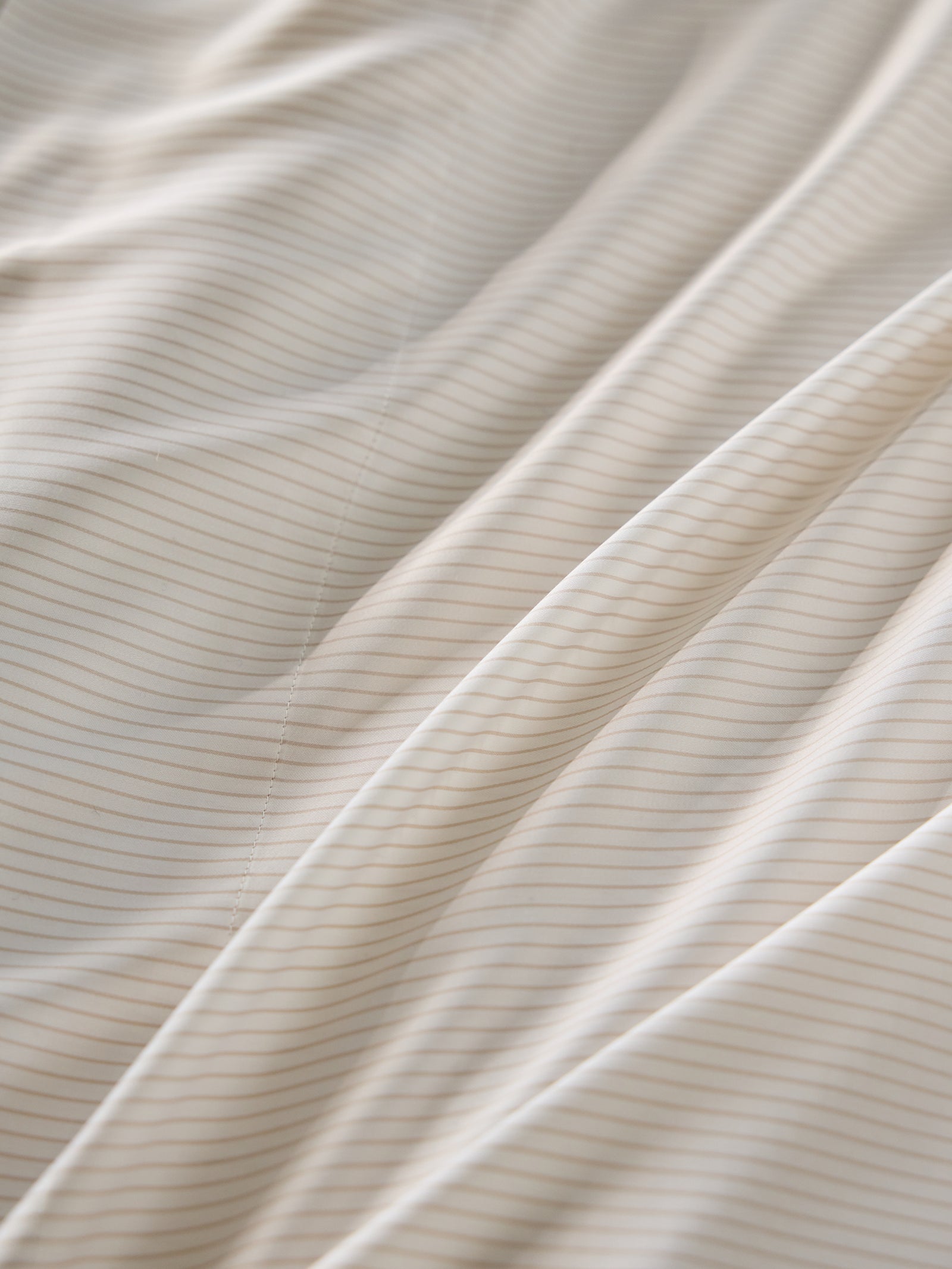 A close-up of Cozy Earth's Bamboo Fitted Sheet reveals its smooth, beige surface with subtle vertical pinstripes. The lighting creates soft shadows and highlights, accentuating the sheet's texture and gentle folds. 