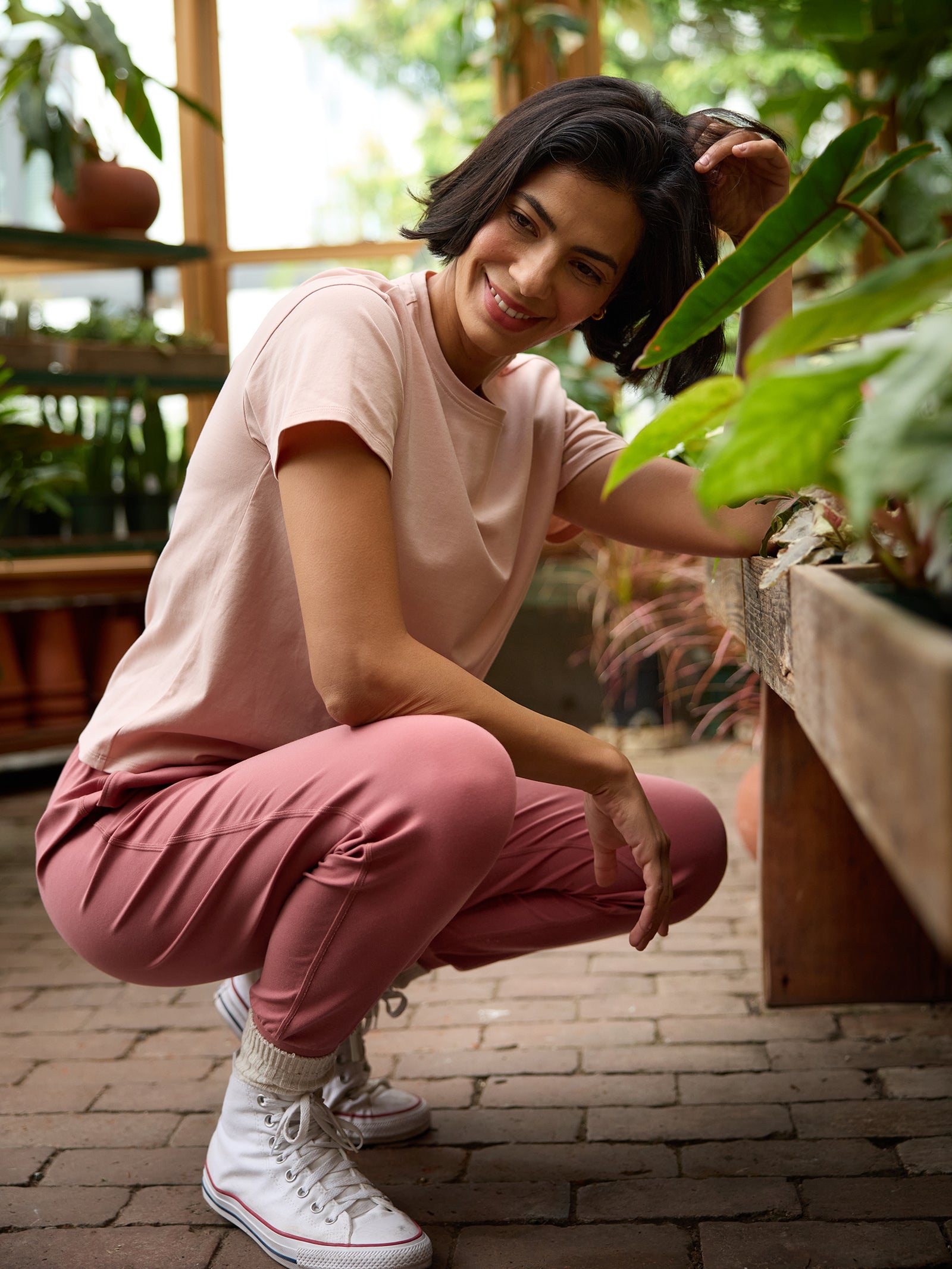 A person with short dark hair, dressed in Cozy Earth's Women's All Day Tee in light pink, along with matching pink pants and white sneakers, kneels beside a wooden planter box filled with green plants in a greenhouse. Smiling as they adjust a plant leaf, they are surrounded by an assortment of potted plants. 