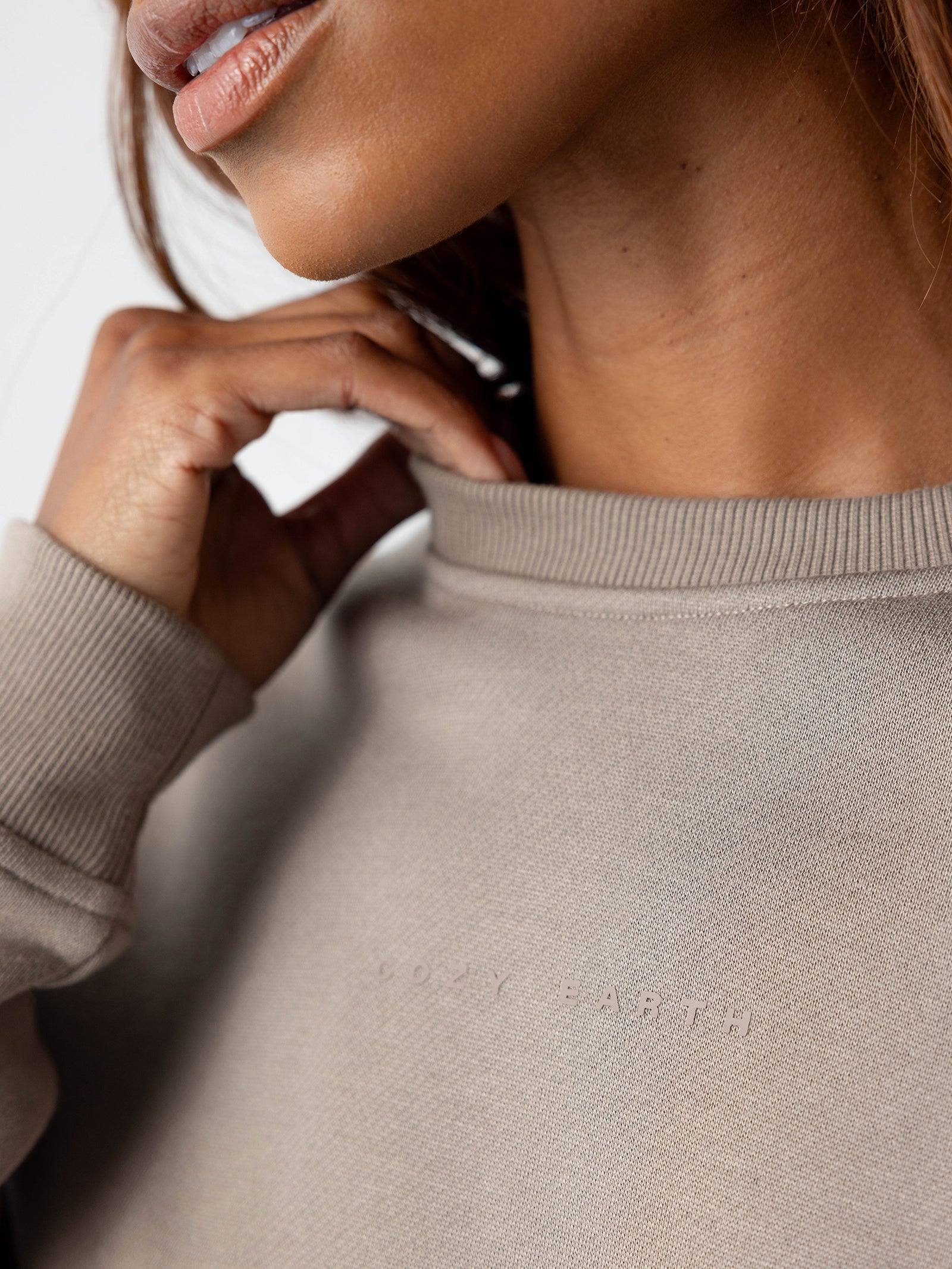 Close-up of a person wearing the Women's CityScape Crewneck in light grey, with "Cozy Earth" subtly embroidered across the chest. The person's hand is gently touching the neckline, and part of their face and neck is visible. 