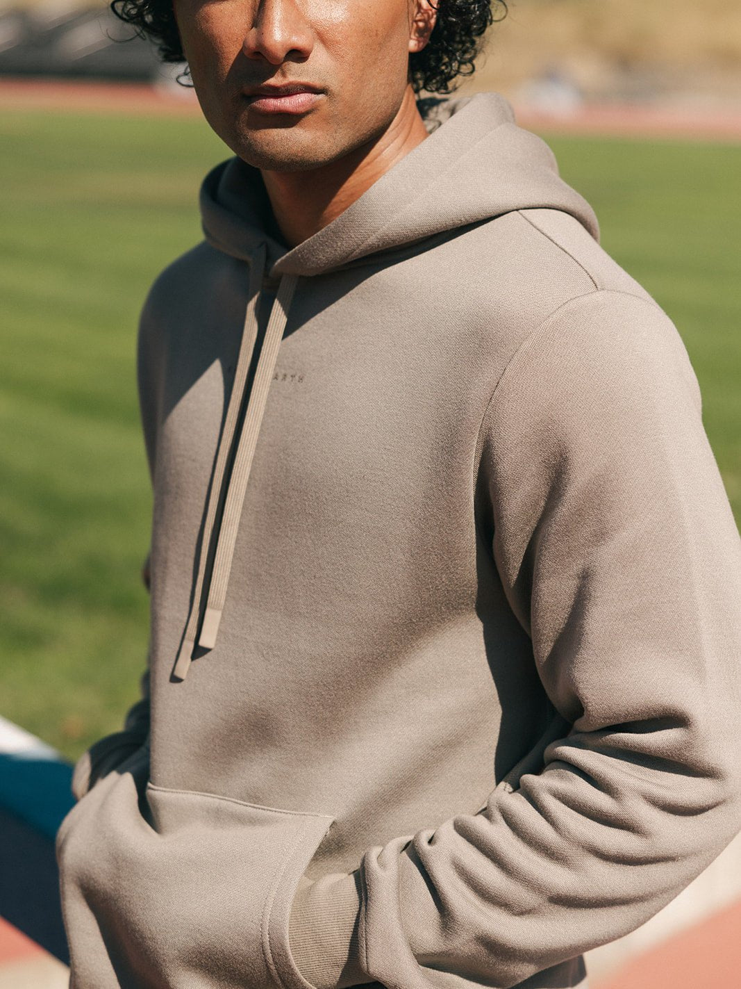 A person with curly hair stands outside on a sunny day wearing a beige Men's CityScape Hoodie by Cozy Earth, with their hands in the front pockets. The background is a blurred field and walking track. 