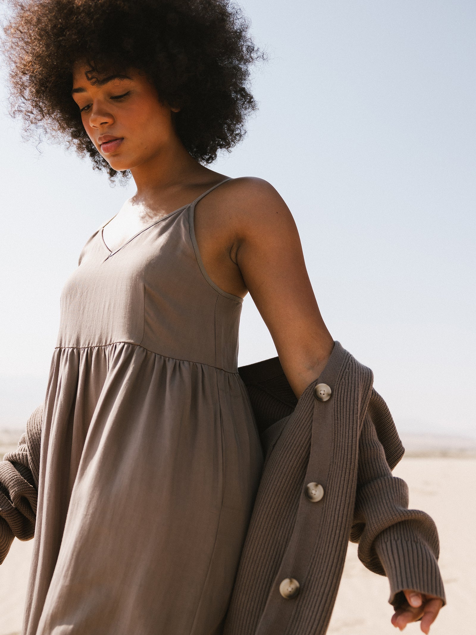 A person with curly hair stands outdoors under a clear blue sky, wearing a sleeveless beige dress and an oversized classic cardigan by Cozy Earth, casually draped off one shoulder. 