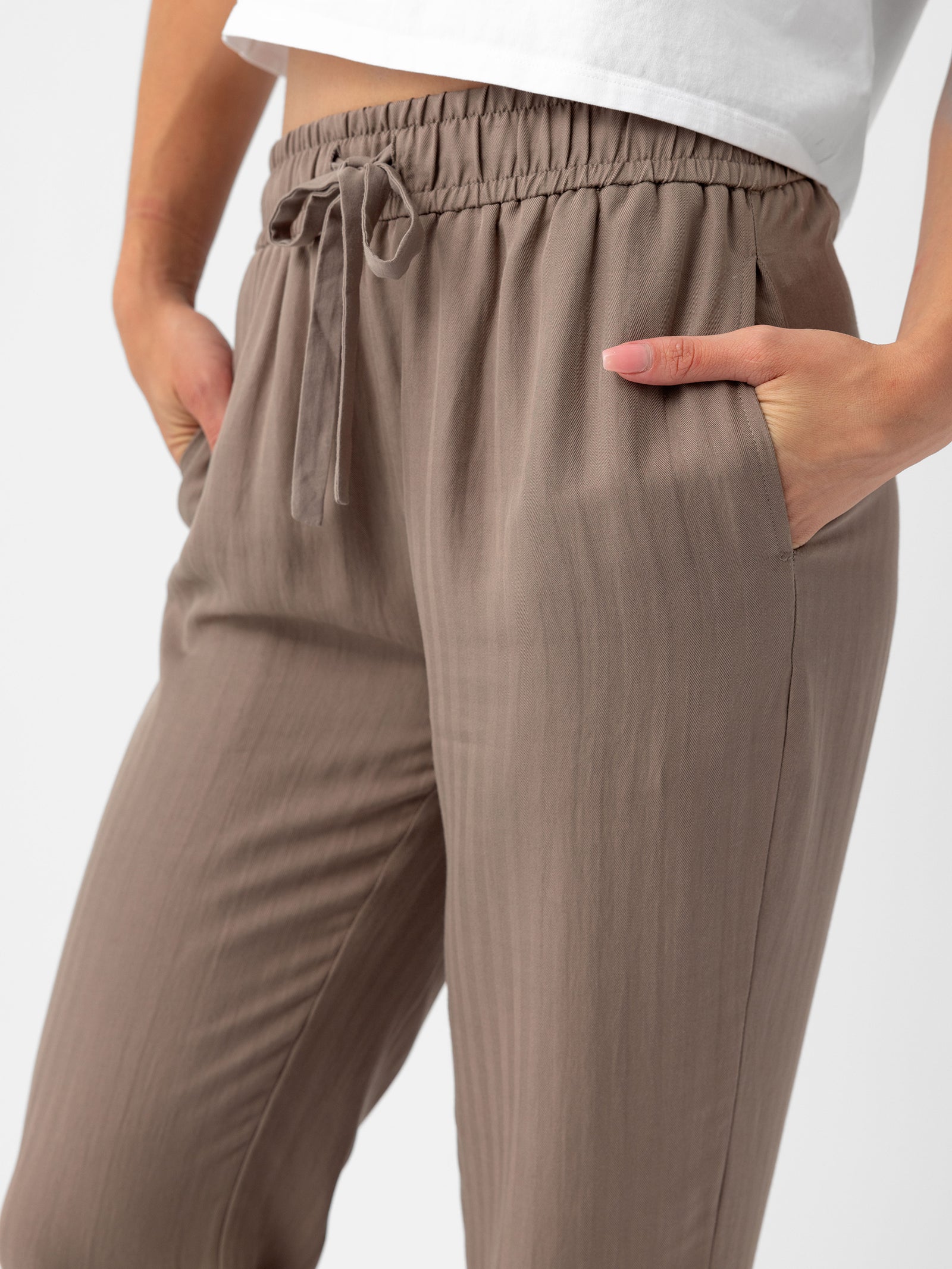 A person is shown from the waist down wearing Cozy Earth Women's Sunset Cropped Pant in a taupe color, with their hands in the pockets. They are also wearing a white t-shirt. The background is plain white. 