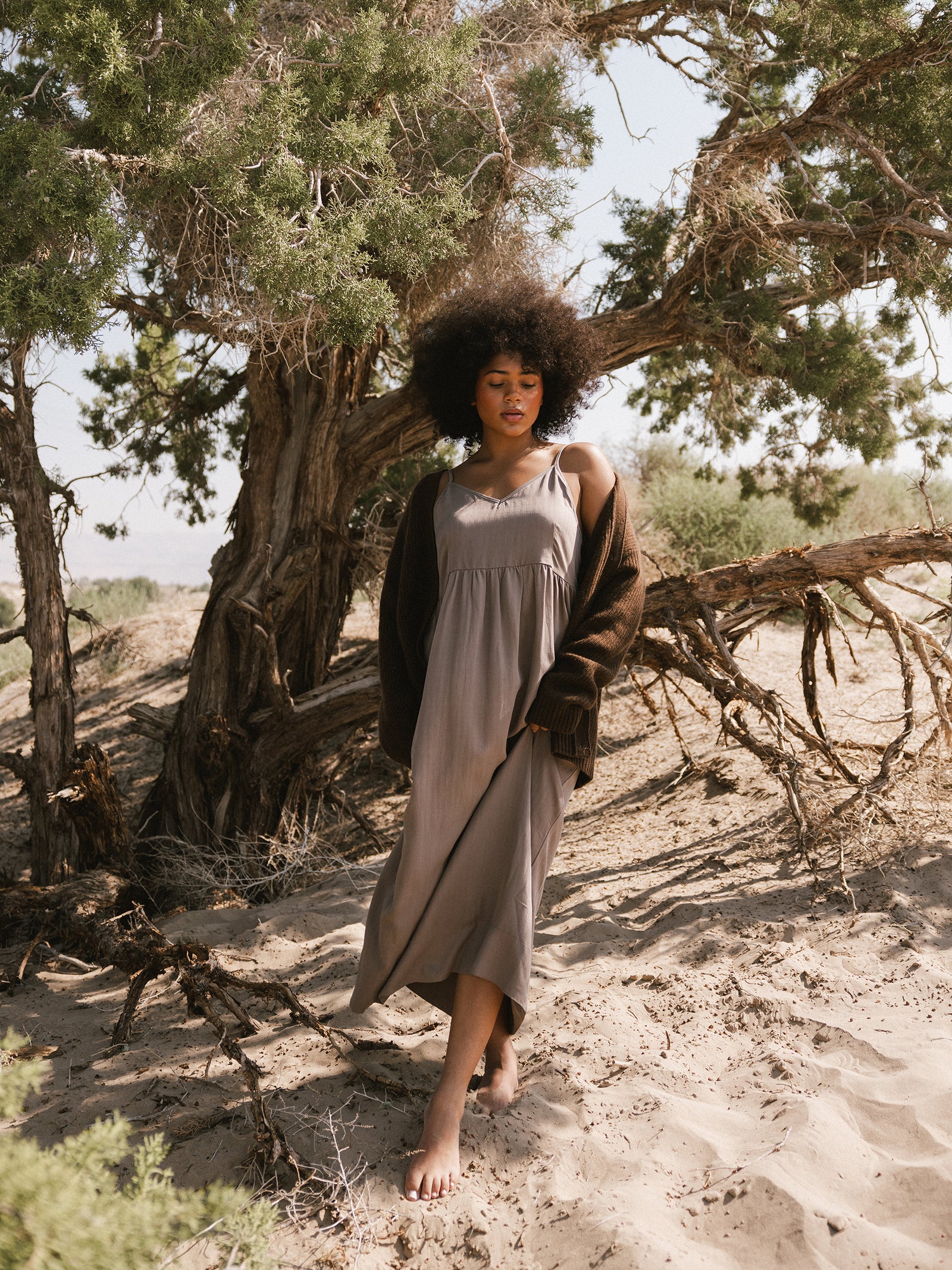 A person with a voluminous afro hairstyle walks barefoot in a sandy, wooded area. They are wearing the long, flowing Women's Sunset Gathered Dress from Cozy Earth and a brown cardigan. The background features a large, gnarled tree and sparse vegetation under a clear sky. 