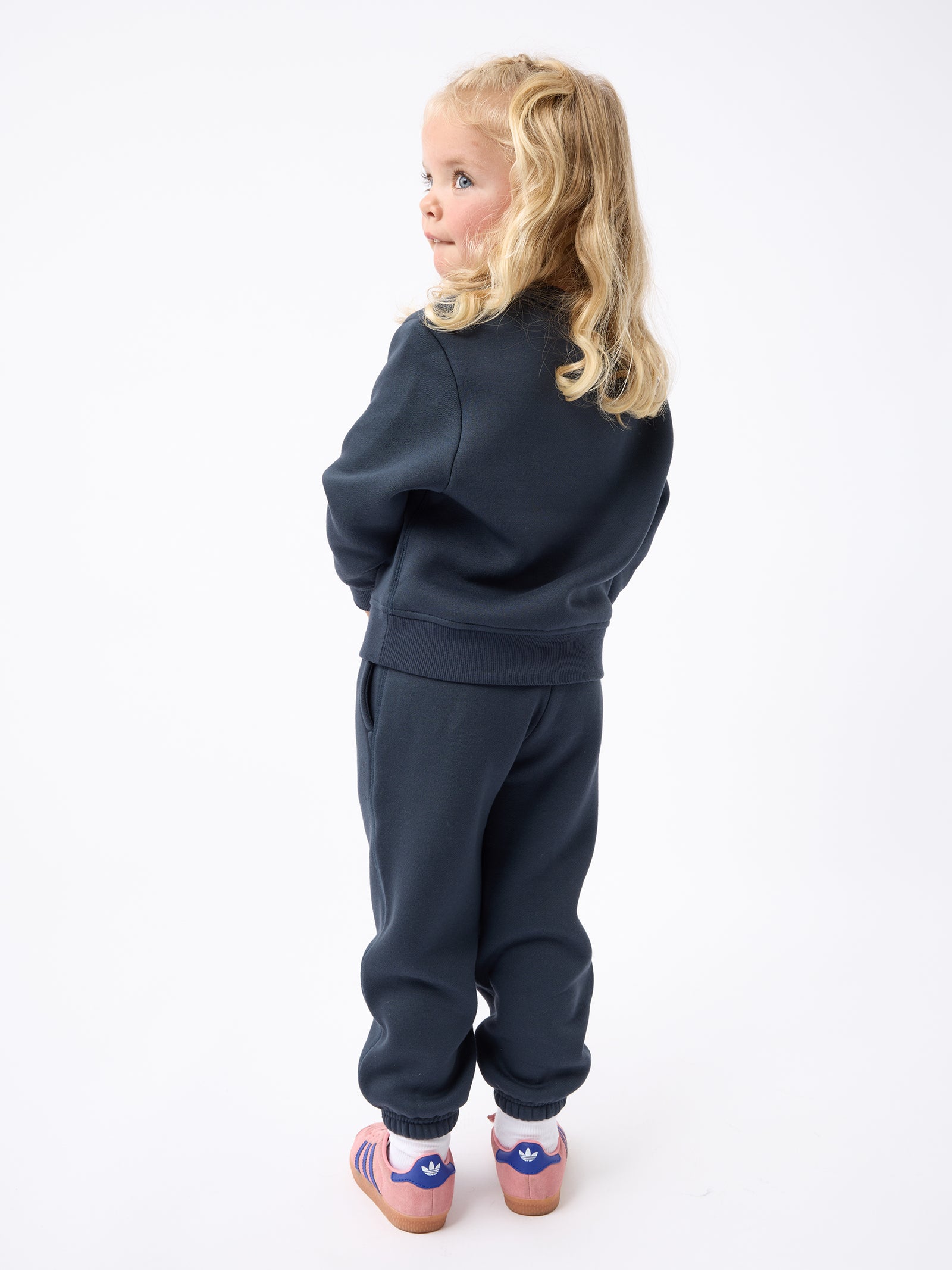 A young child with long blonde hair, viewed from the back and slightly turned to the left, is wearing a Cozy Earth Kid's CityScape Crewneck in dark blue paired with pink striped sandals. The child stands against a plain white backdrop. 