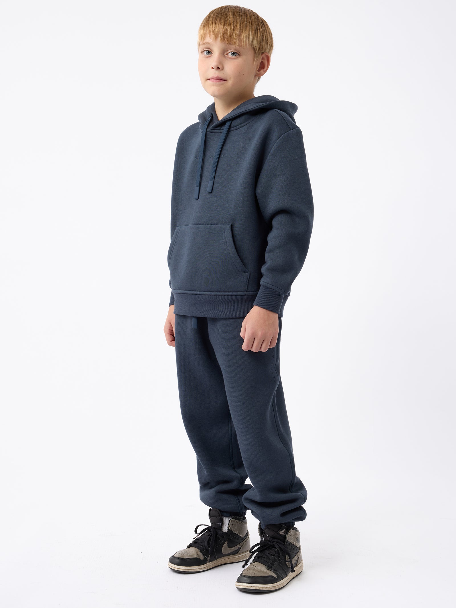 A young boy stands against a plain white background, dressed in the Kid's CityScape Hoodie by Cozy Earth, paired with matching navy blue sweatpants. He is wearing black and white sneakers and has his hands comfortably tucked into the hoodie pockets, gazing slightly to the side. 