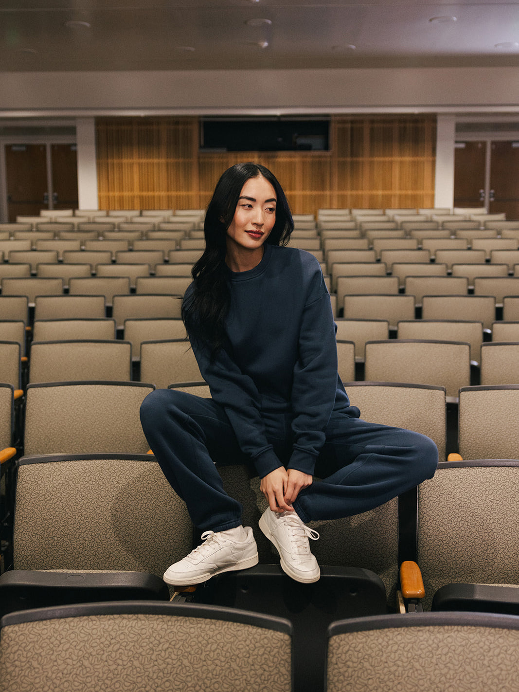 A person with long dark hair is sitting casually on the backrest of a seat in an empty auditorium. They are wearing a matching navy blue Women's CityScape Crewneck from Cozy Earth and white sneakers, with both feet resting on the seat in front of them. The setting is spacious and well-lit. 