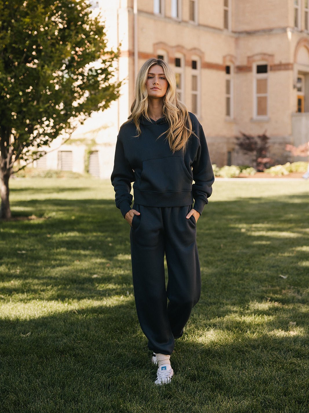 A person with long blonde hair stands on a grassy area in front of a building with brick and stone walls. They are wearing the Women's CityScape Cropped Hoodie by Cozy Earth, paired with dark-colored sweatpants and white sneakers. With their hands in their pockets, they look directly at the camera. A tree is partially visible on the left. 