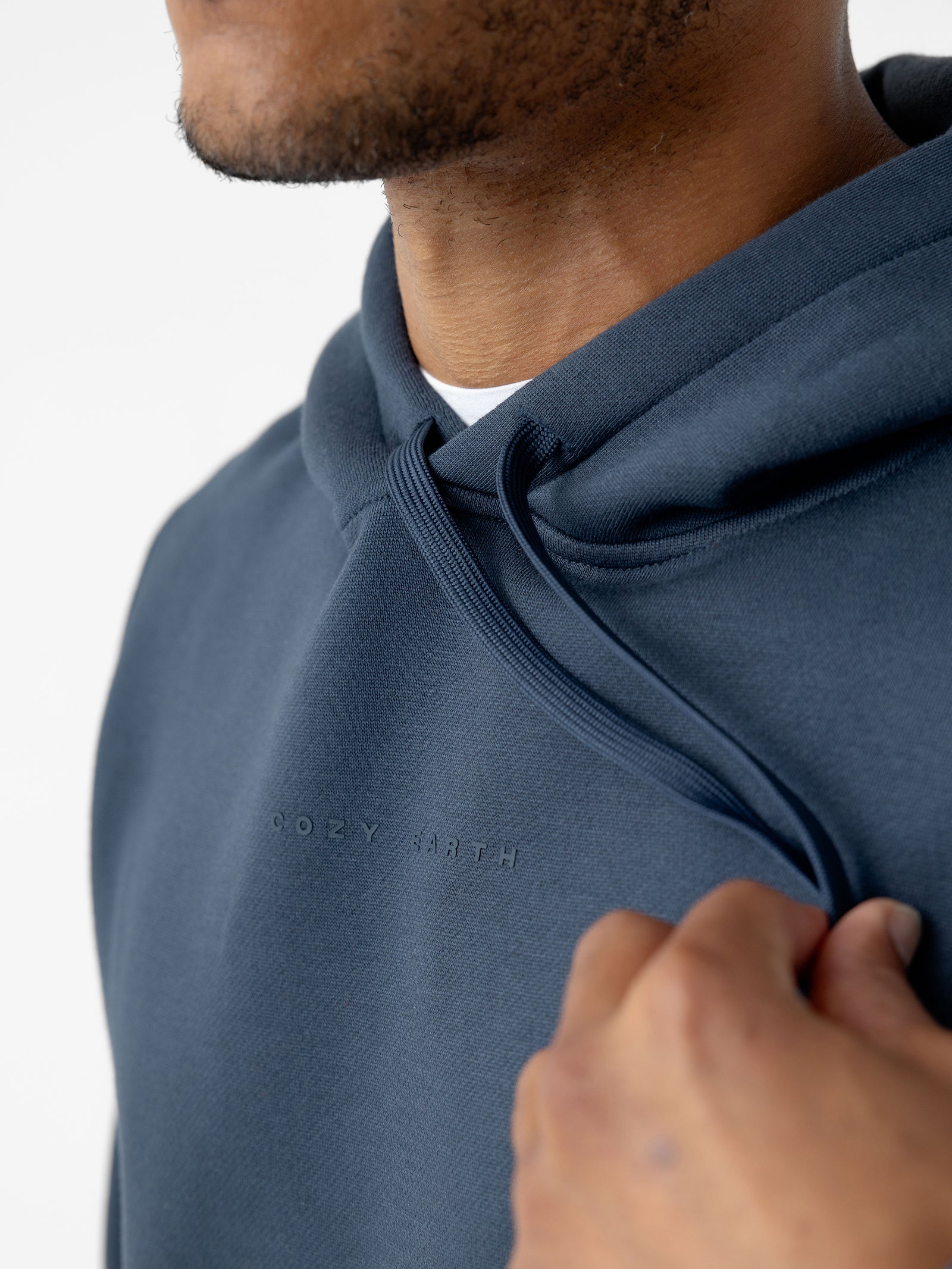 Close-up of a person wearing a Men's CityScape Hoodie in dark blue by Cozy Earth, with "COZY EARTH" embossed on the chest. The hoodie's drawstrings are visible, and the person is partially shown from the side, holding the drawstring with their right hand. The background is white. 