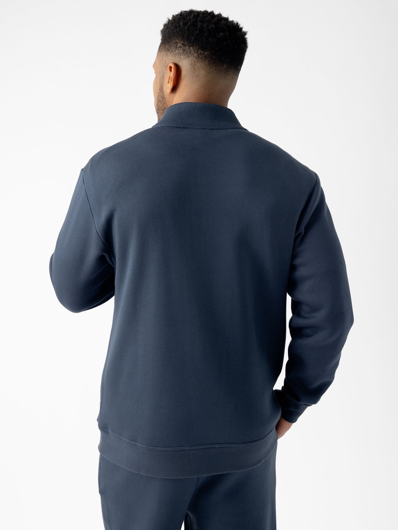 A person with short, curly hair is standing facing away from the camera, wearing a dark blue Men's CityScape Quarter Zip and matching pants from Cozy Earth. The background is plain white. 