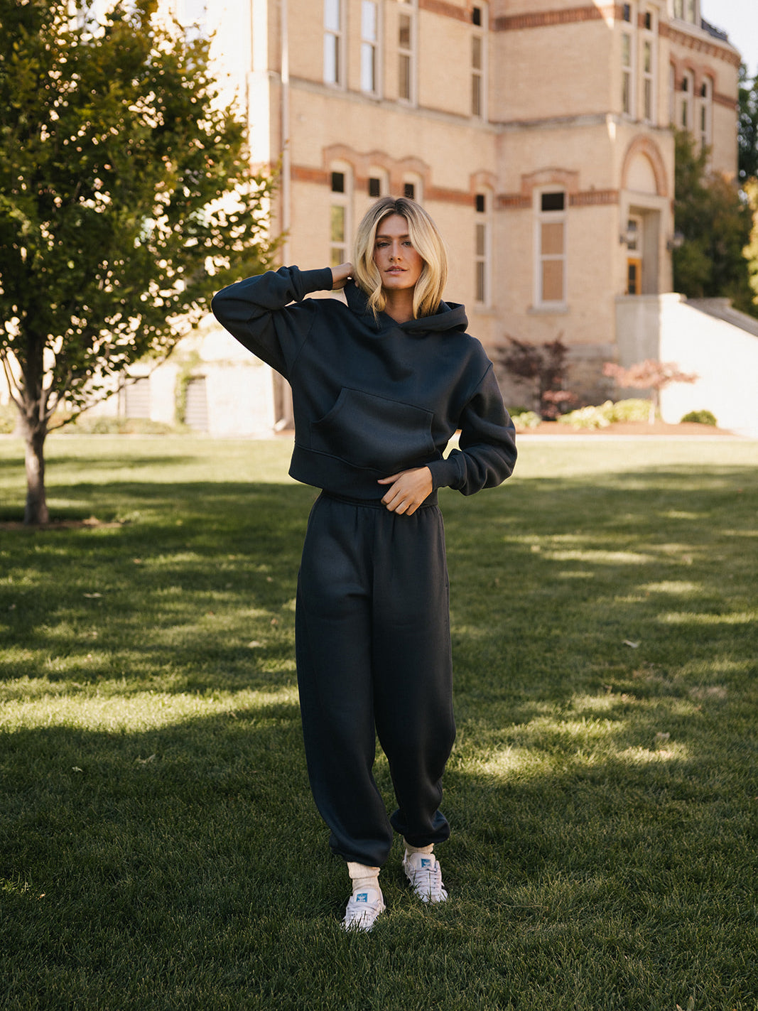 A woman stands on a grassy lawn in front of a large brick building, wearing a dark hoodie and matching Cozy Earth Women's CityScape Sweatpant. She has one hand raised to her head, and her expression is neutral. There are trees and shrubbery around the area. 