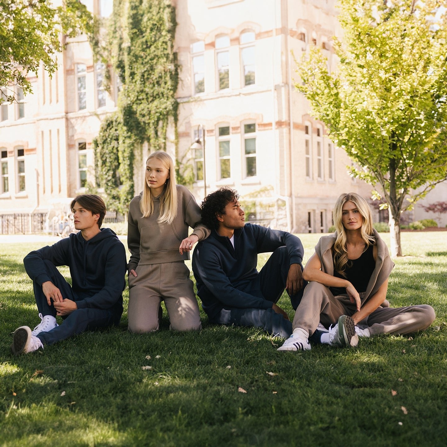 Four people sit on the grass outdoors, all dressed in casual and comfortable attire. Among them, one individual wears the Men's CityScape Sweatpant from Cozy Earth. In the background, there's a large light-colored building with windows and trees, suggesting the scene is set in a park or university campus. 