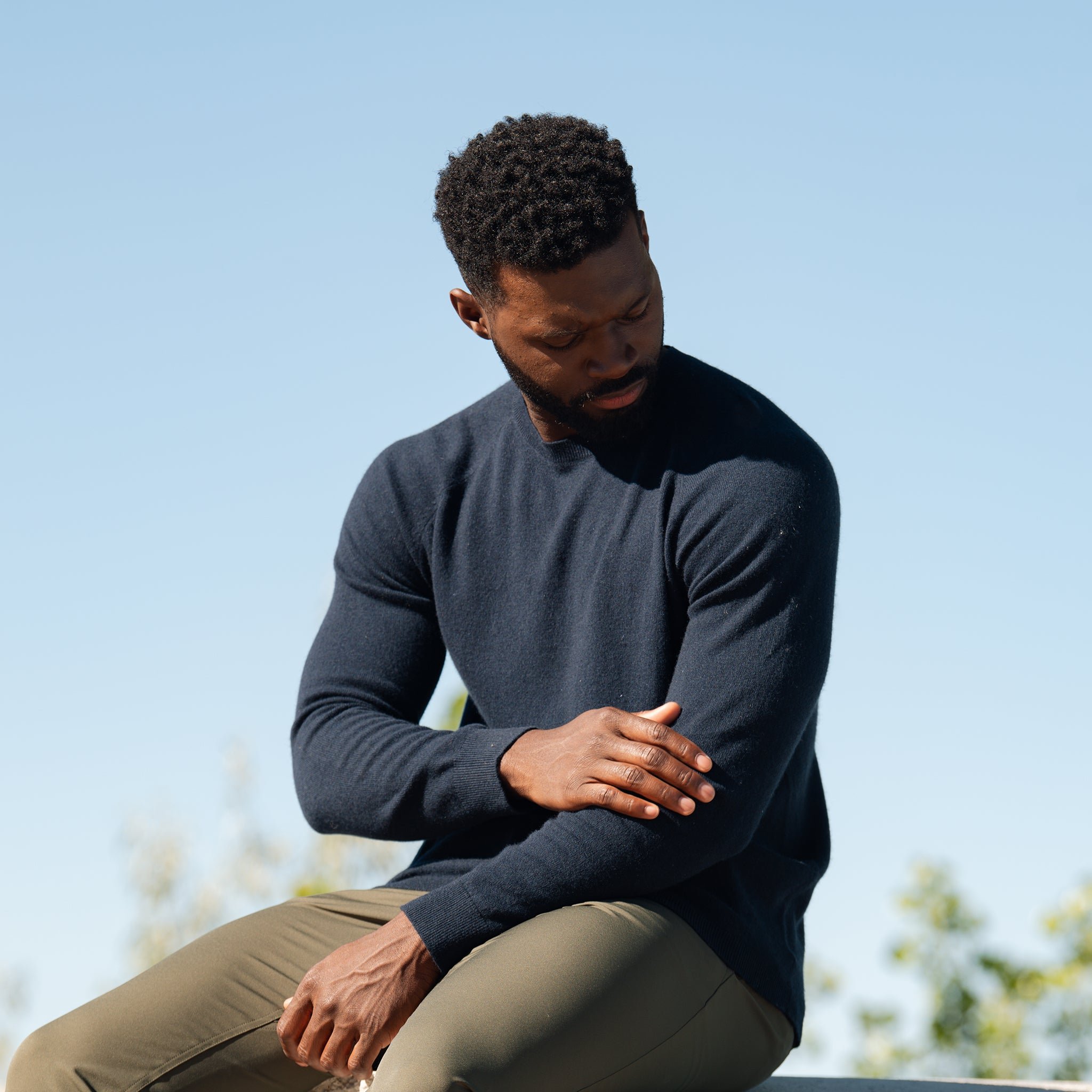 A person sitting outdoors on a sunny day, looking down at their arm with one hand touching the opposite elbow. They are wearing a navy blue Men's Crewneck Sweater by Cozy Earth and olive green pants. The background shows a clear blue sky with some blurred greenery. 