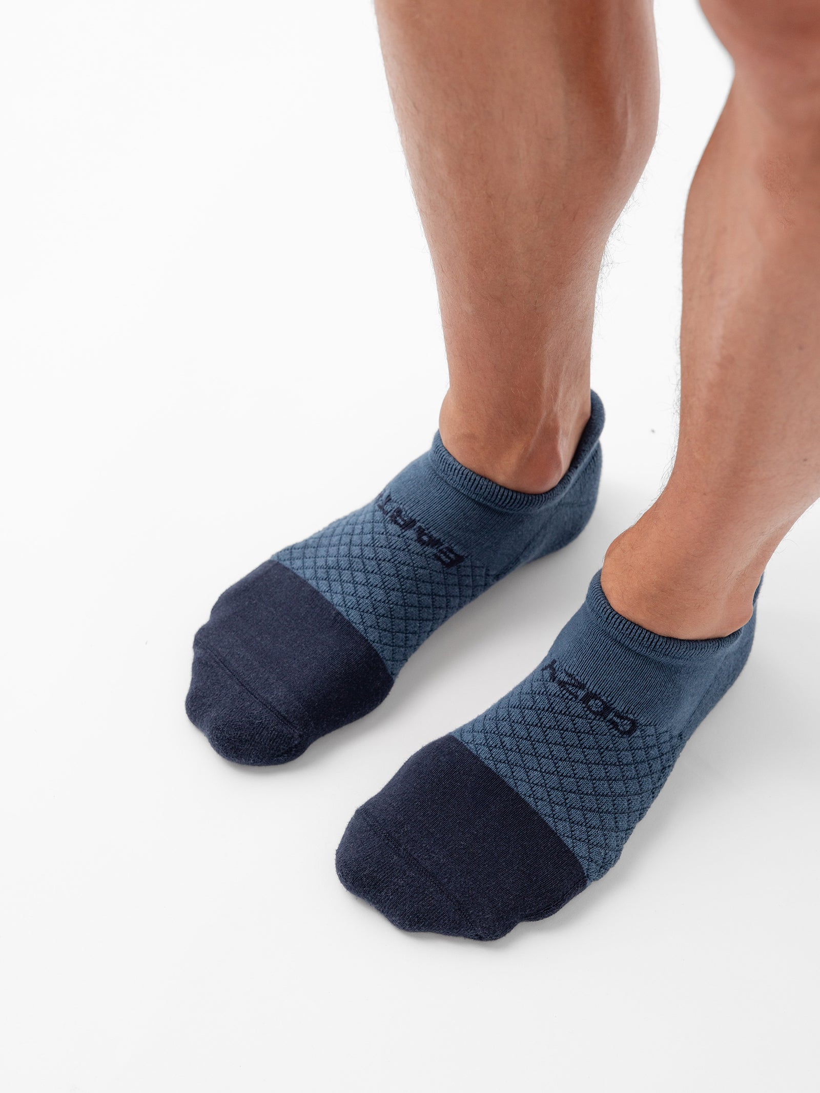 A person is wearing a pair of Essential Ankle Socks from Cozy Earth. The socks feature a low-cut design with dark blue toes and heels, complemented by a lighter blue middle section adorned with a diamond pattern. The person is standing on a white surface, displaying the full view of their calves and feet. 