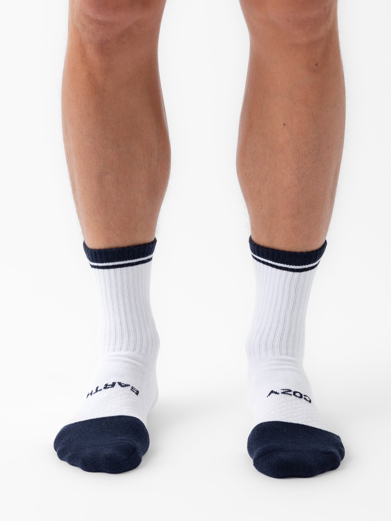 A person is wearing Cozy Earth's Essential Calf Sock 2-Pack, featuring white socks with black toes and heels, along with black stripes at the top. The left sock displays the word "EARTH" near the toe, while the right sock features "AEON." They are standing on a white surface. 