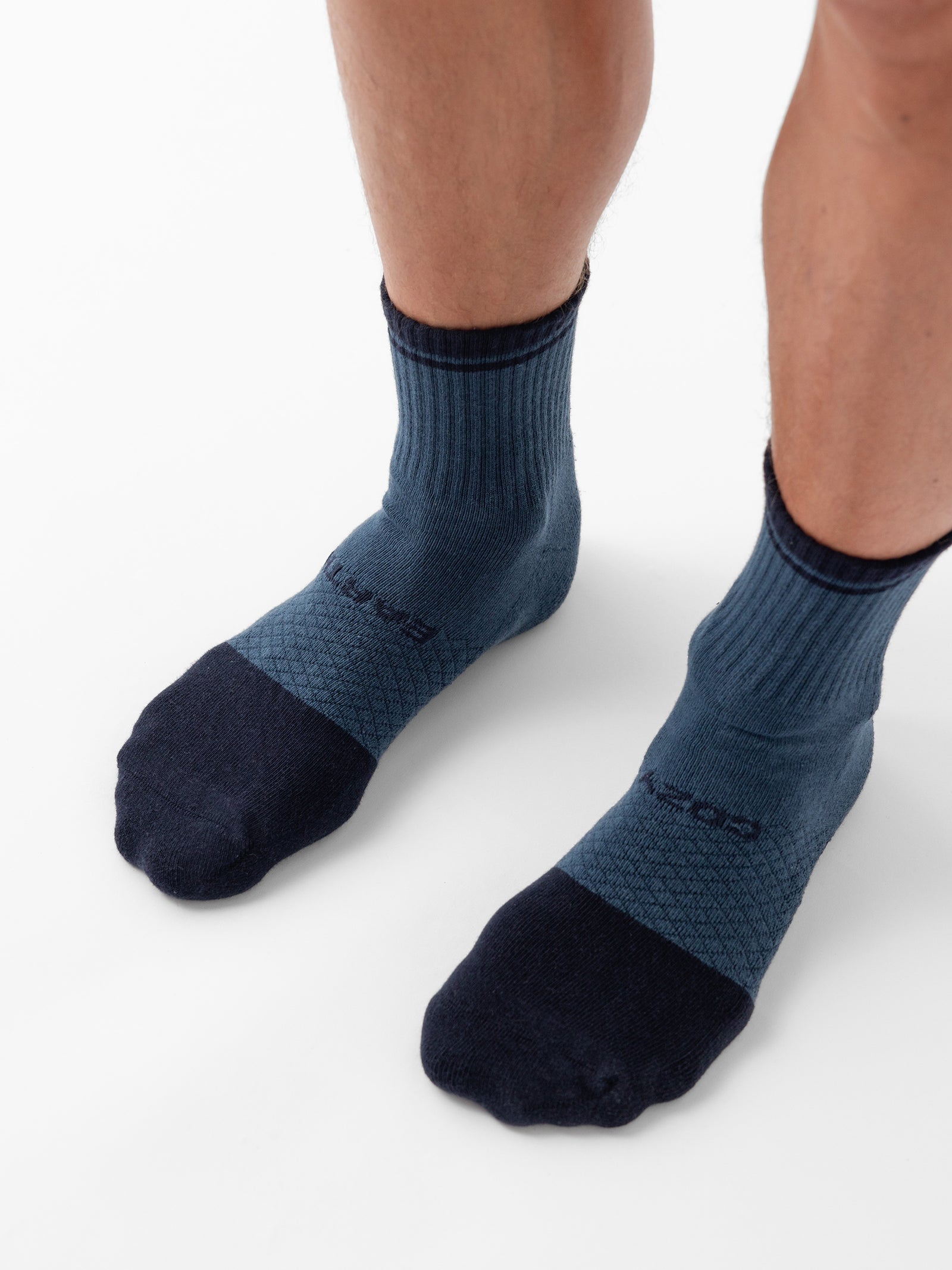 Close-up of a person's lower legs and feet wearing two-tone Essential Quarter Socks from Cozy Earth. The socks feature dark blue at the toes and heels, transitioning to a lighter blue covering the rest of the foot and ankle, with a ribbed texture extending up the ankle. The person stands on a white background. 