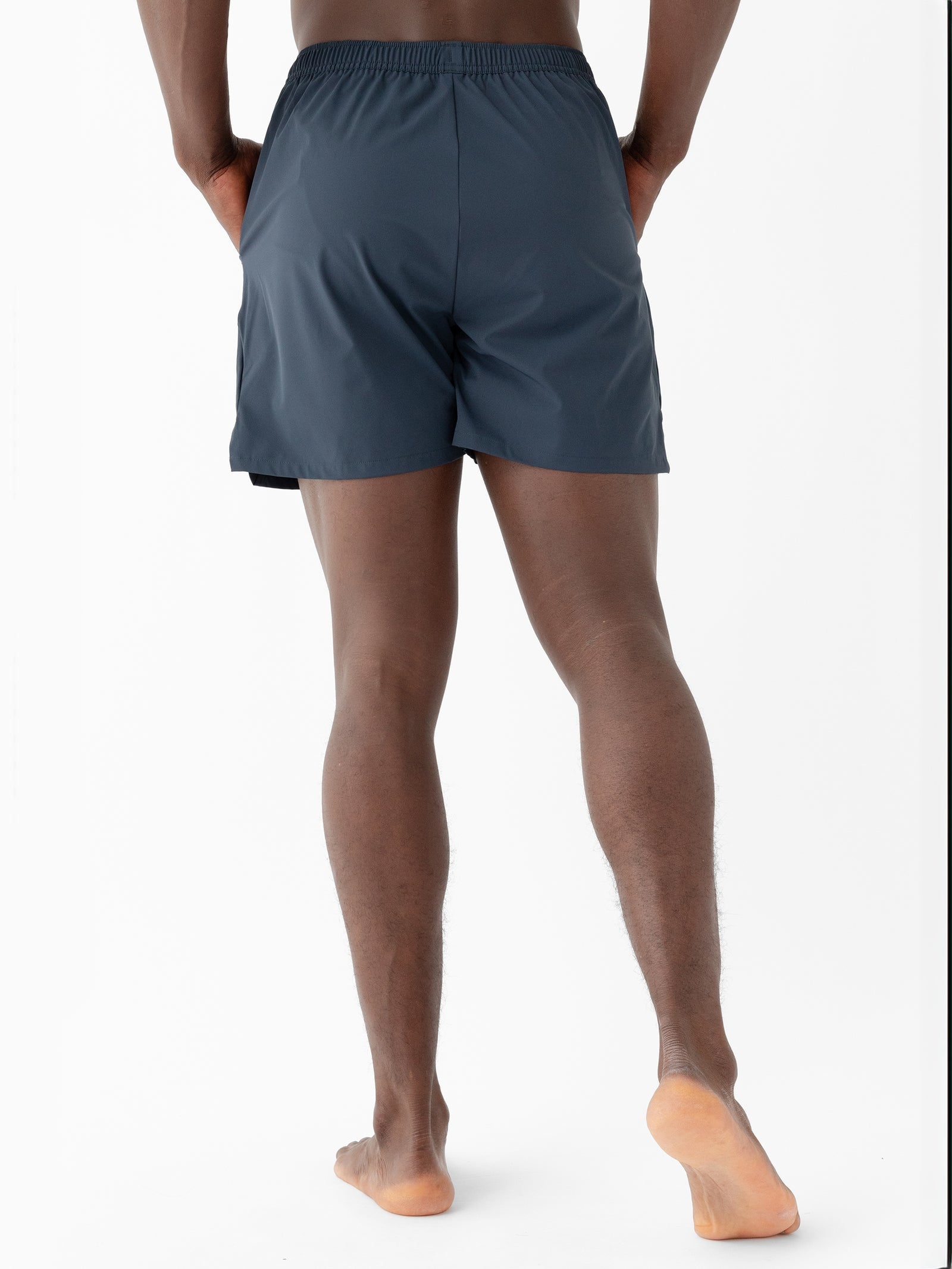 Rear view of a person standing barefoot against a white background. They are wearing dark-colored, above-the-knee Men's Performance Sleep Shorts by Cozy Earth with hands in pockets and feet slightly apart. The fabric appears lightweight and suitable for sports or active wear. 