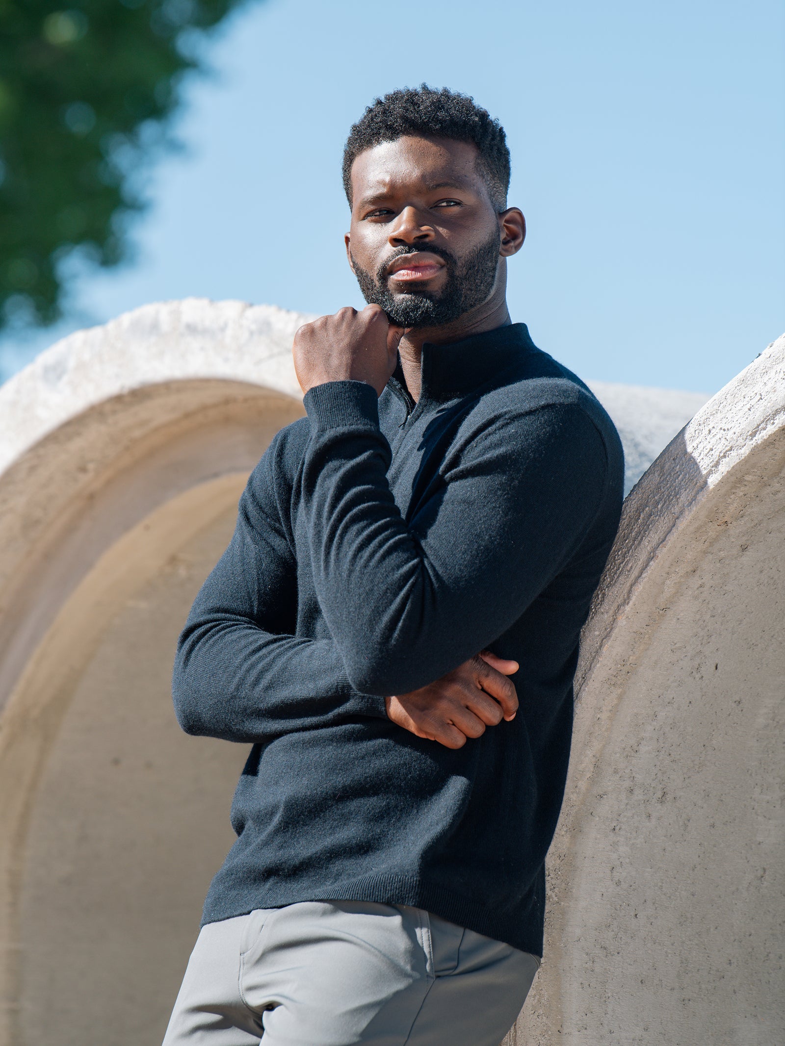 A man with a beard and short hair stands outdoors, leaning against a large stone structure. He is wearing a dark Cozy Earth Men's Quarter Zip Sweater and light pants, with one hand resting on his chin. The background features greenery and a clear blue sky. 