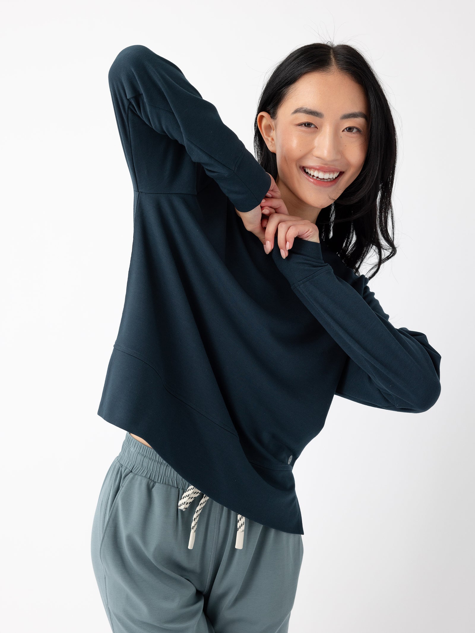 A woman with long, dark hair is smiling and posing in a playful manner. She is wearing the Women's StudioLite Crewneck by Cozy Earth, paired with light-colored pants featuring a drawstring waist. The background is plain white. 