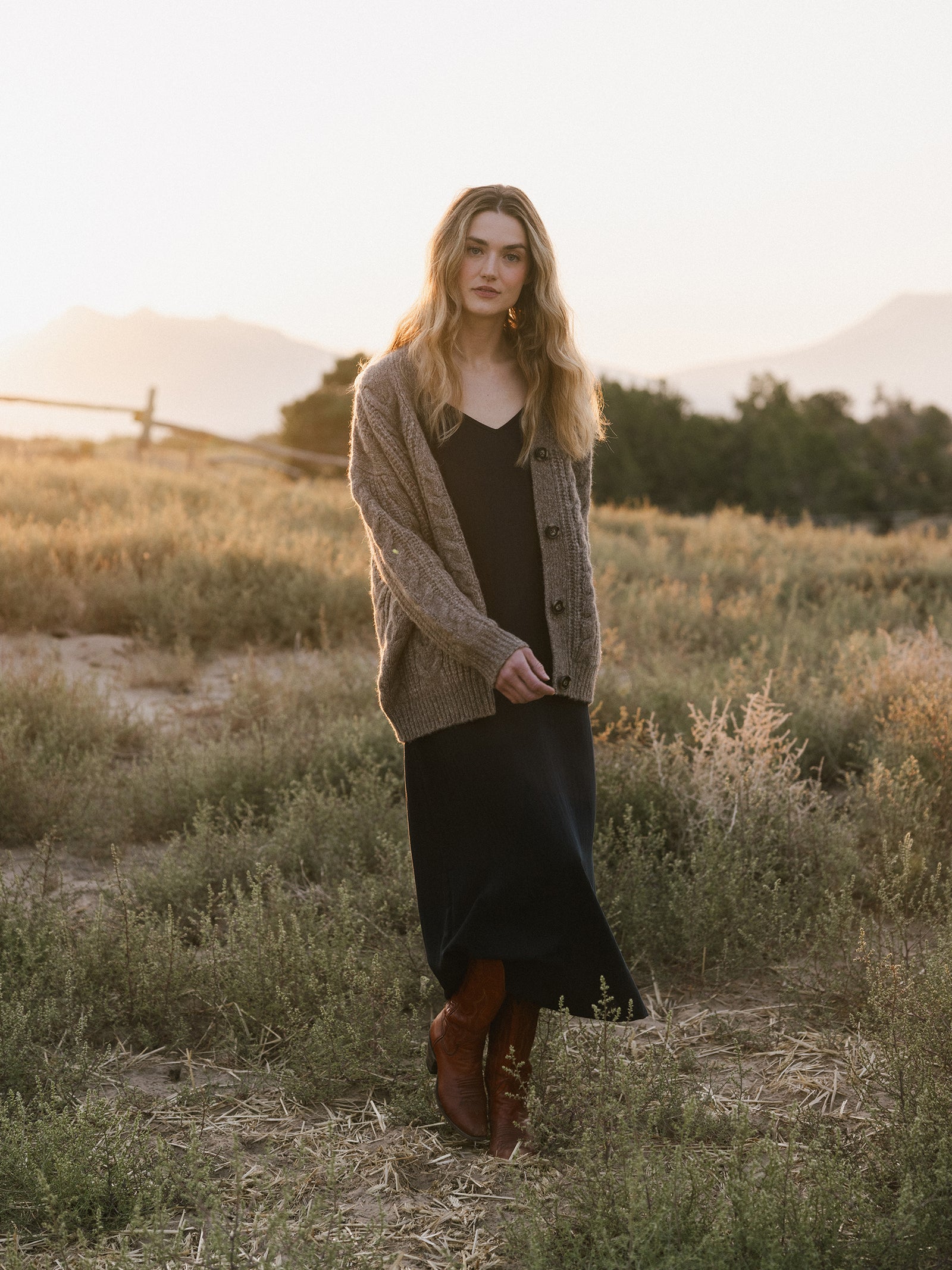 A woman with long, wavy hair stands in an open field at sunset. She is wearing Cozy Earth's Women's Sunset Slip Dress paired with a chunky knit cardigan and brown boots. A wooden fence and trees are visible in the background, along with distant mountains. 