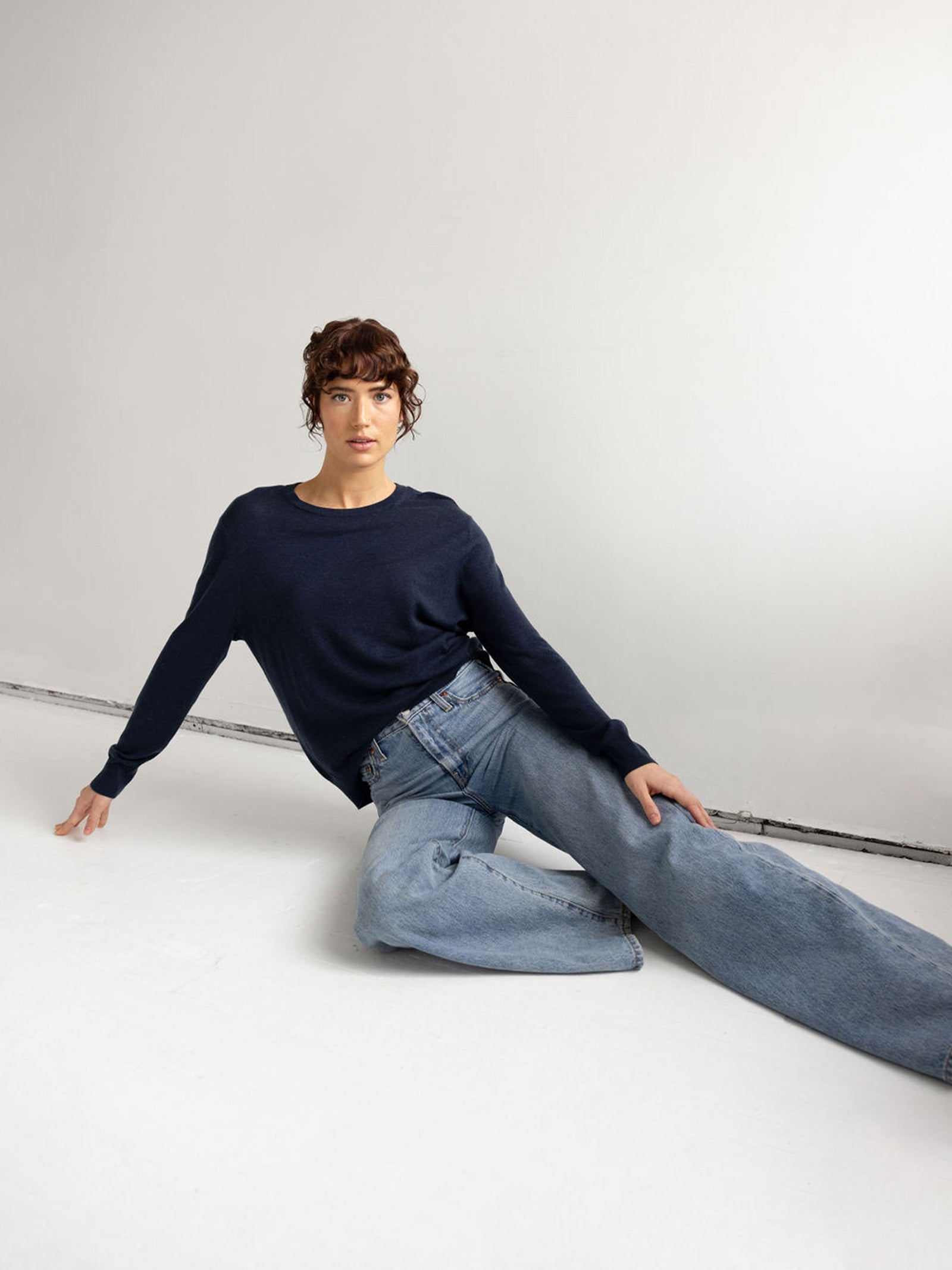 Woman lying on her side wearing jeans and an eclipse airknit sweater with white background 