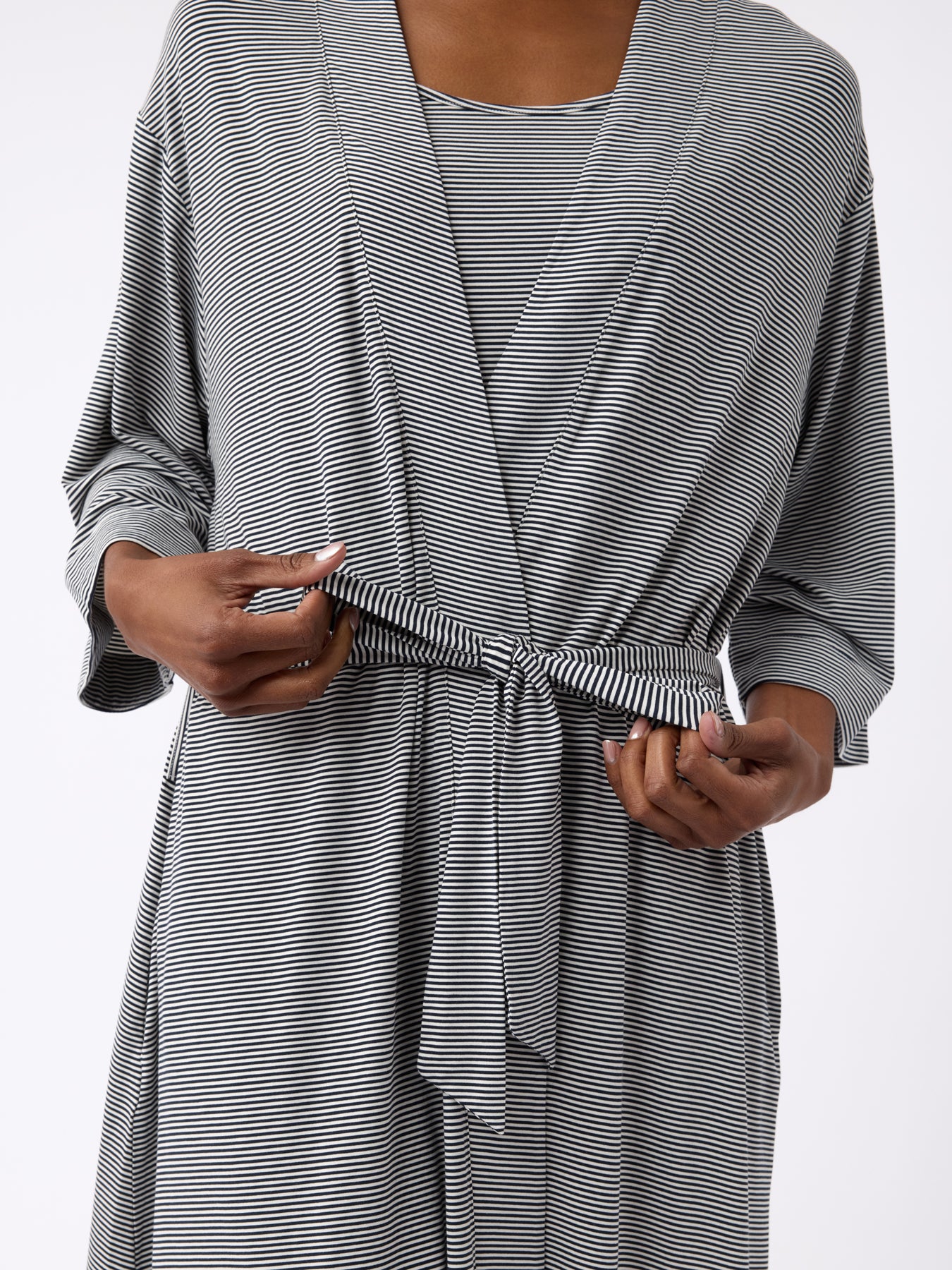 A person adjusts the waist tie of a black and white striped Cozy Earth Women's Bamboo Stretch-Knit Kimono Robe, featuring three-quarter sleeves and a wrap design, with an image centered on their torso and hands. 