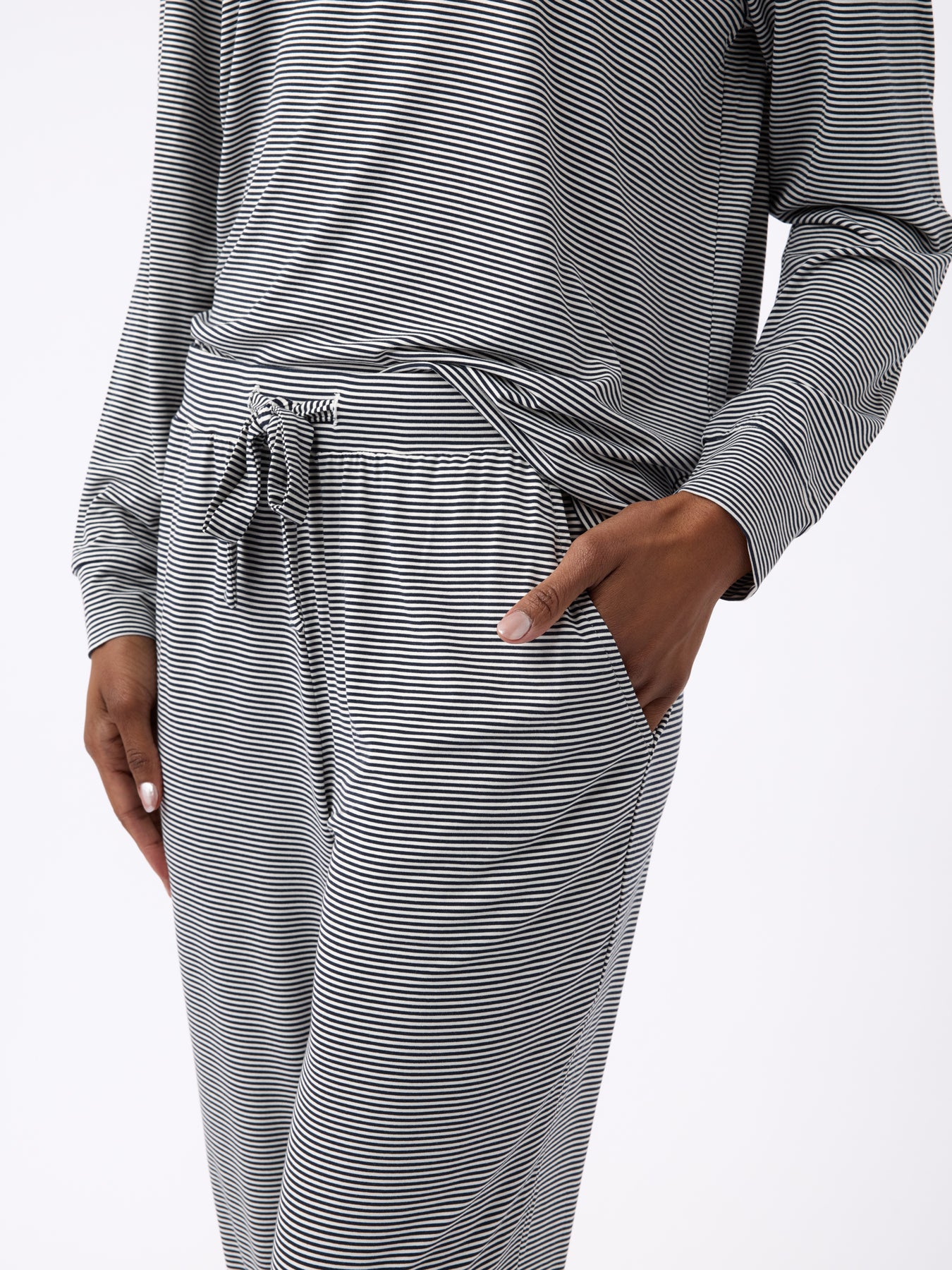 A person is wearing Cozy Earth's Women's Bamboo Stretch-Knit Tapered Pant, featuring long sleeves, black and white stripes, and a tied waist detail. They're standing in a casual pose with one hand in their pocket against a white background. 