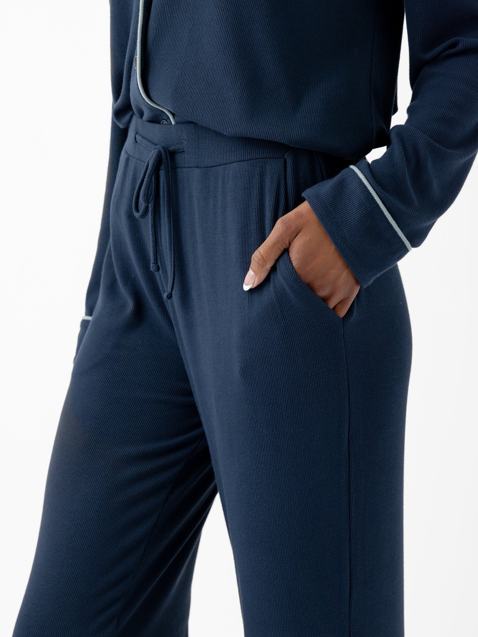 A person wearing Cozy Earth's Women's Bamboo Rib Knit Classic Pajama Pant in navy blue with white trim is shown from the chest down. The person has their right hand in the pocket of the pants, showcasing neatly manicured nails. The background is plain white. 