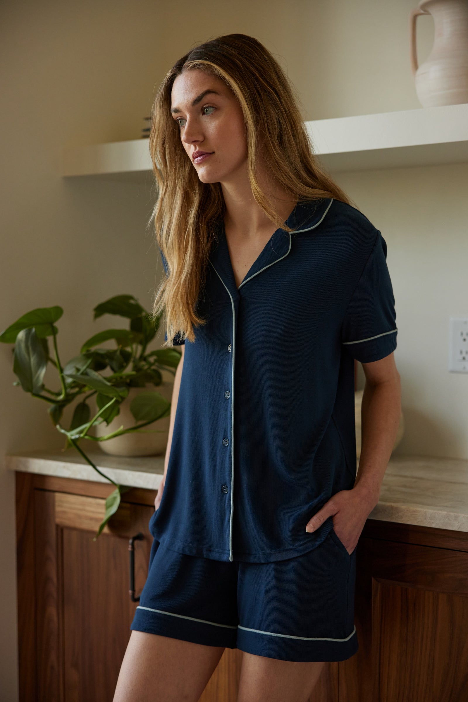 A person with long, wavy hair is standing in a kitchen, wearing Cozy Earth's blue Women's Bamboo Rib Knit Classic Short Sleeve Pajama Top and matching shorts with white trim. Their hands are in their shorts pockets, and a plant sits on the counter behind them. The background includes a shelf with a vase. 