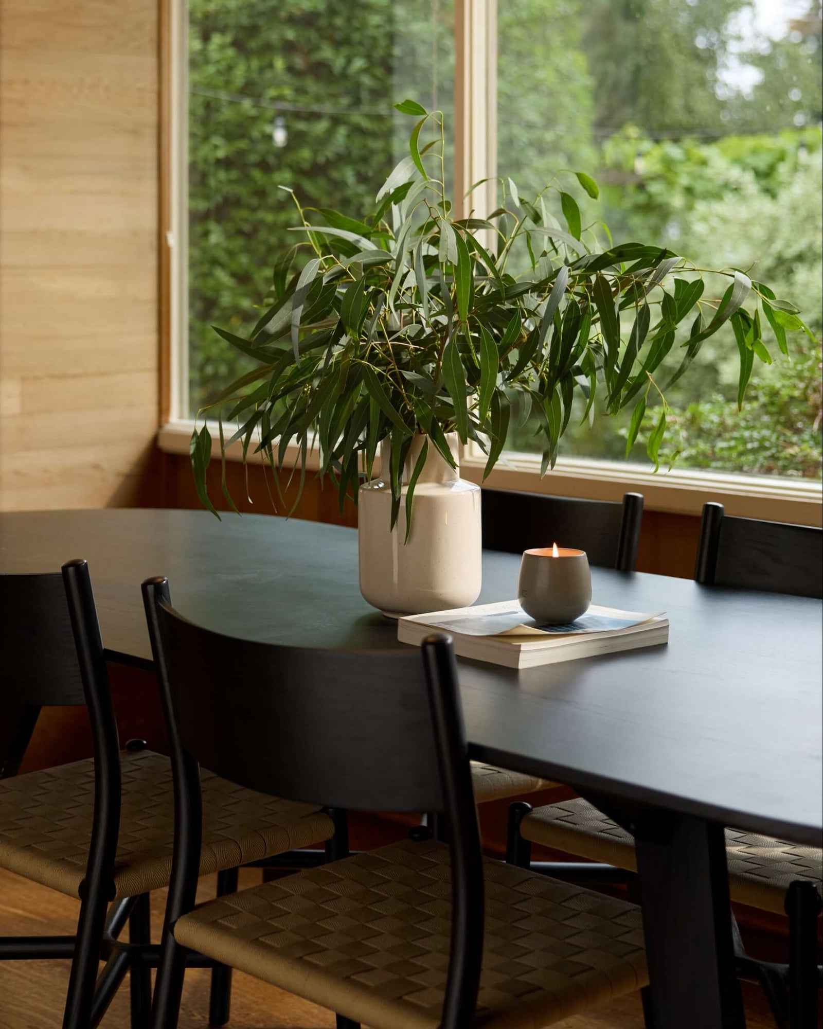 A black dining table with woven-back chairs holds a Cozy Earth Candle EMBER, a white vase of green foliage, and books, set by a large window overlooking lush greenery.