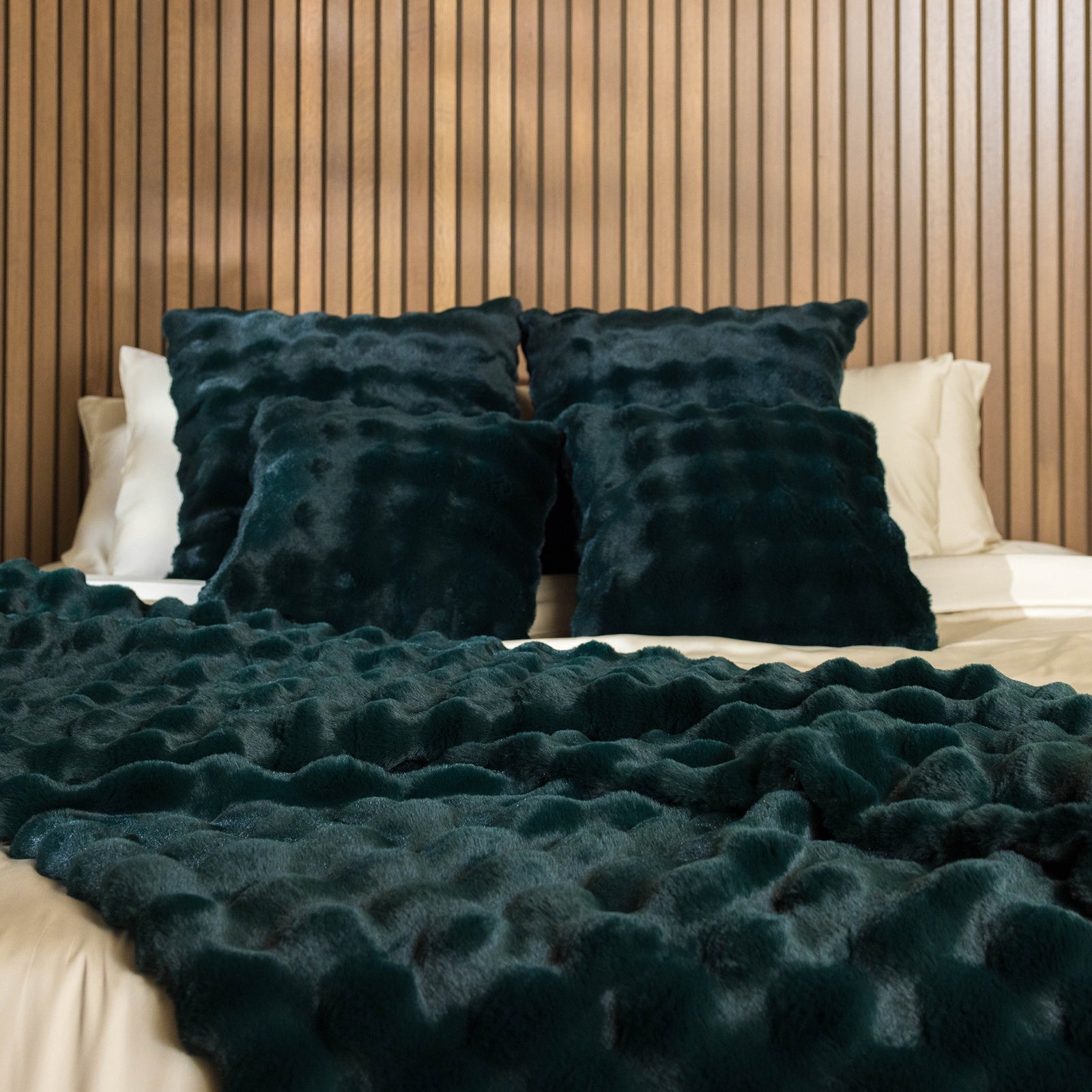 A cozy bed featuring four Bubble Cuddle Pillows in a dark green hue, complemented by a matching throw blanket from Cozy Earth. The bed is set against a backdrop of vertical wooden slats, creating a warm and inviting atmosphere. 