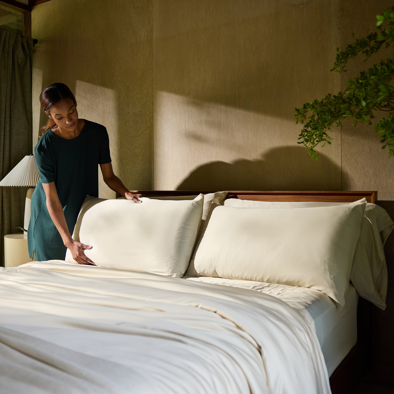 A person in a green dress is making a bed with Cozy Earth's Bamboo Fitted Sheet and large pillows. The bed is positioned against a wooden wall, with a small table and lamp on the left side. Green plants are visible in the foreground on the right side. 