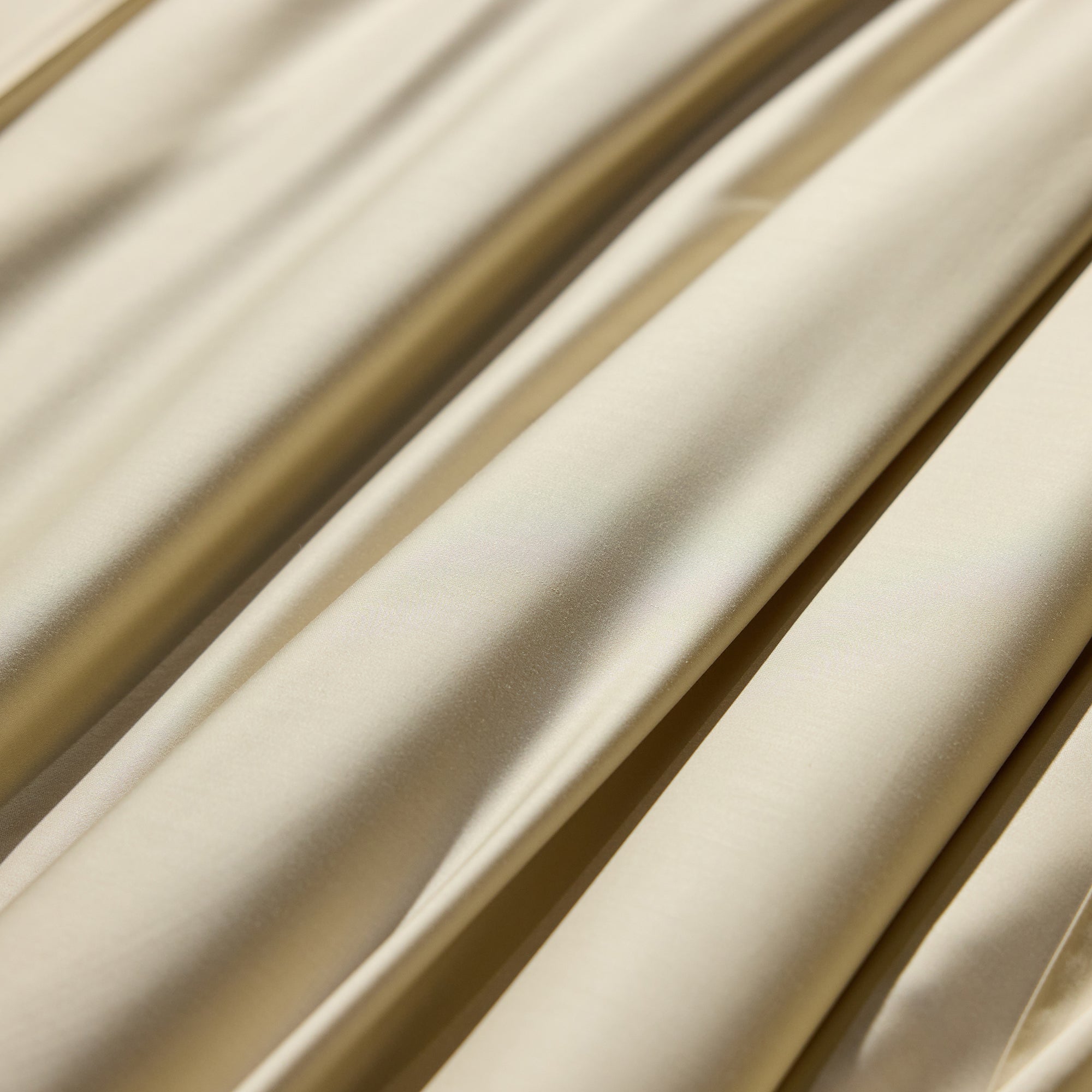 A close-up view of the smooth, shiny fabric of the Bamboo Sheet Set - Wholesale by Cozy Earth, showcasing its beige color with multiple folds. The fabric appears to be silky, reflecting light and displaying variations in texture due to the draping. The folds create a soft, flowing pattern across the image. 