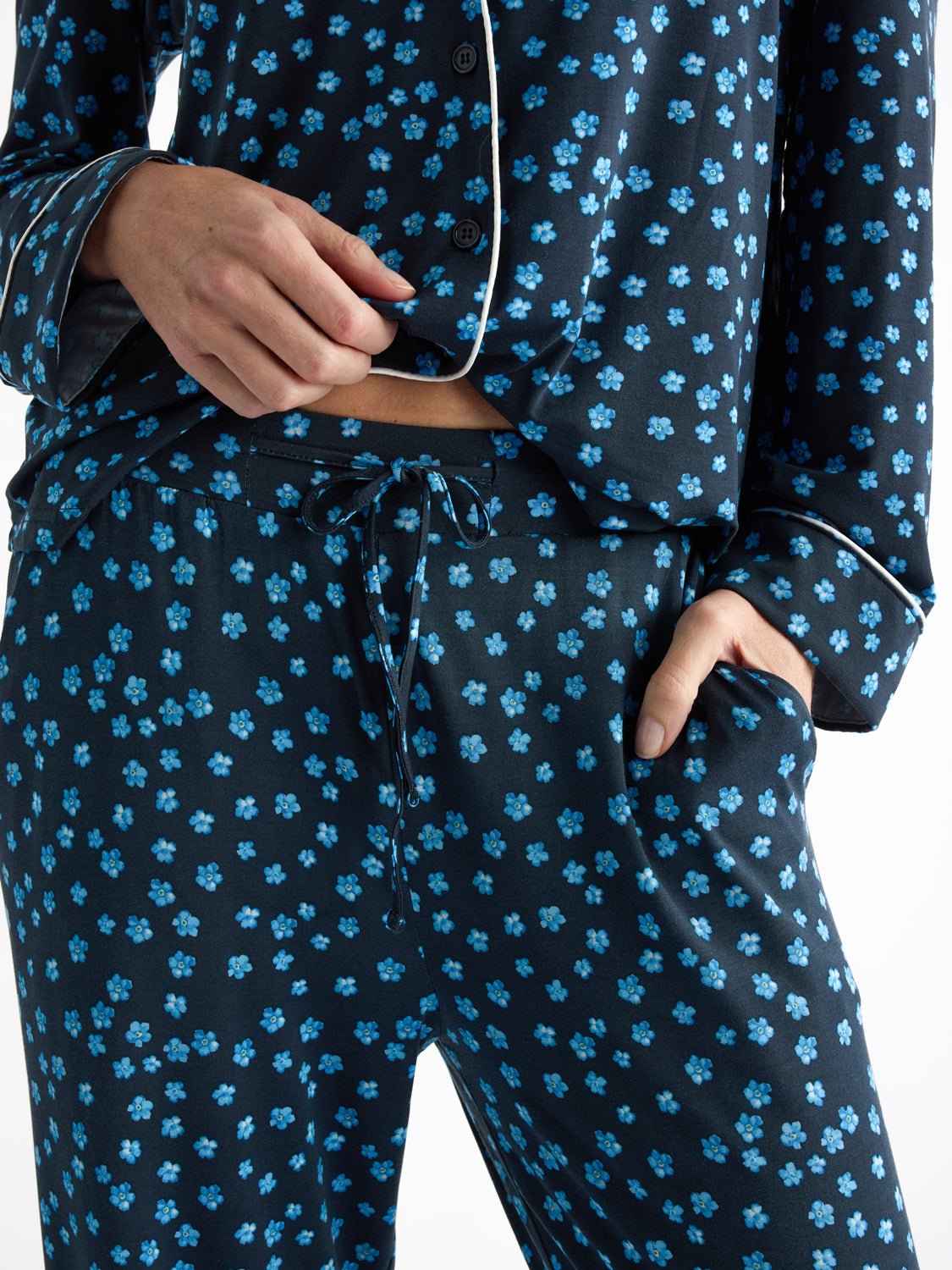 A person is wearing the Women's Bamboo Stretch-Knit Long Sleeve Pajama Set by Cozy Earth, featuring dark tones with bright blue floral patterns. The set includes a buttoned top and drawstring bottoms, with the person's hand in a pocket against a plain white background. |Color:Forget Me Nots