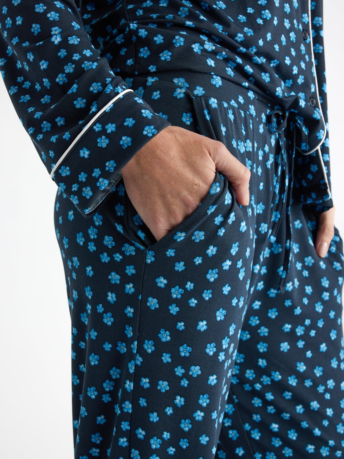 A person wearing Cozy Earth Women's Bamboo Stretch-Knit Pajama Pant in a dark color with a small blue floral pattern. Their right hand rests in a side pocket, highlighting the cuff detail with white piping. The image focuses on the lower torso and arm. 