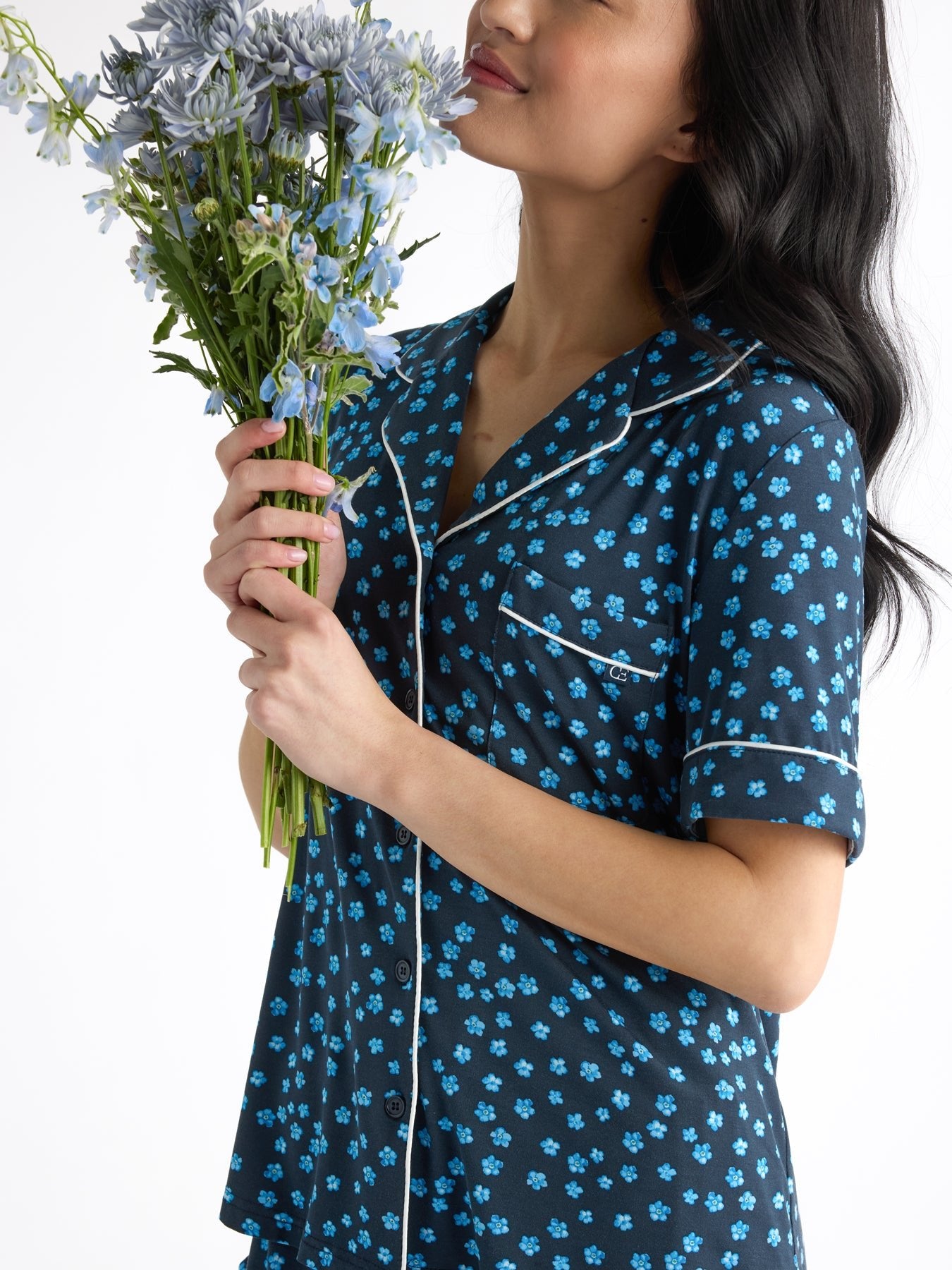 Holding a bouquet of purple and blue flowers, a person with long dark hair wears the Women's Bamboo Stretch-Knit Short Sleeve Pajama Top by HIDE in navy with blue floral patterns and white piping. The background is plain white. 