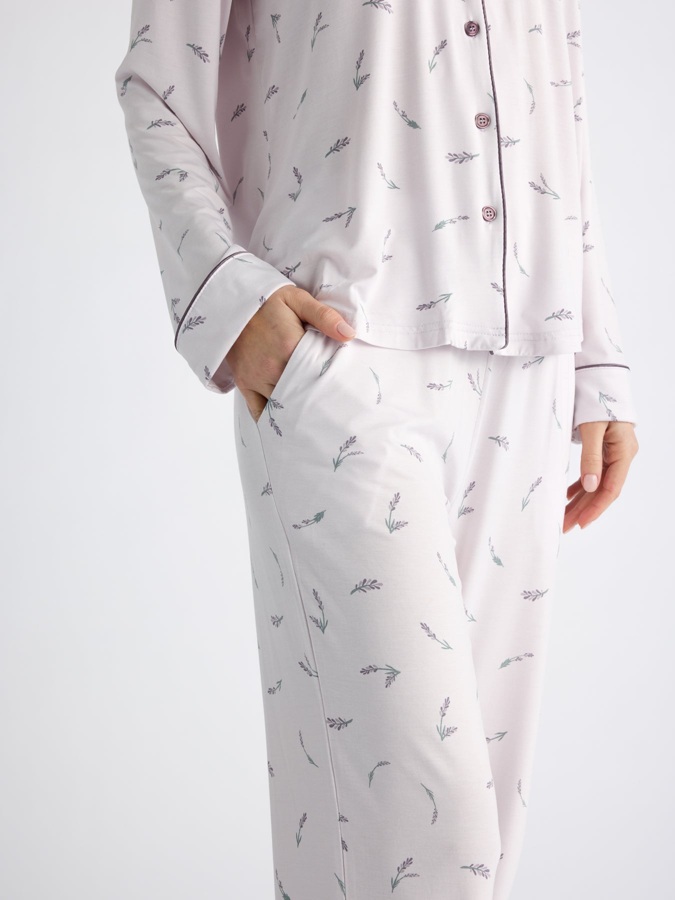 A person is wearing Cozy Earth's Women's Bamboo Stretch-Knit Long Sleeve Pajama Set, featuring cozy, light-colored fabric with a subtle leaf pattern. Their hand rests casually in the pocket of the pants, and the top has buttons. The background is plain and light. 