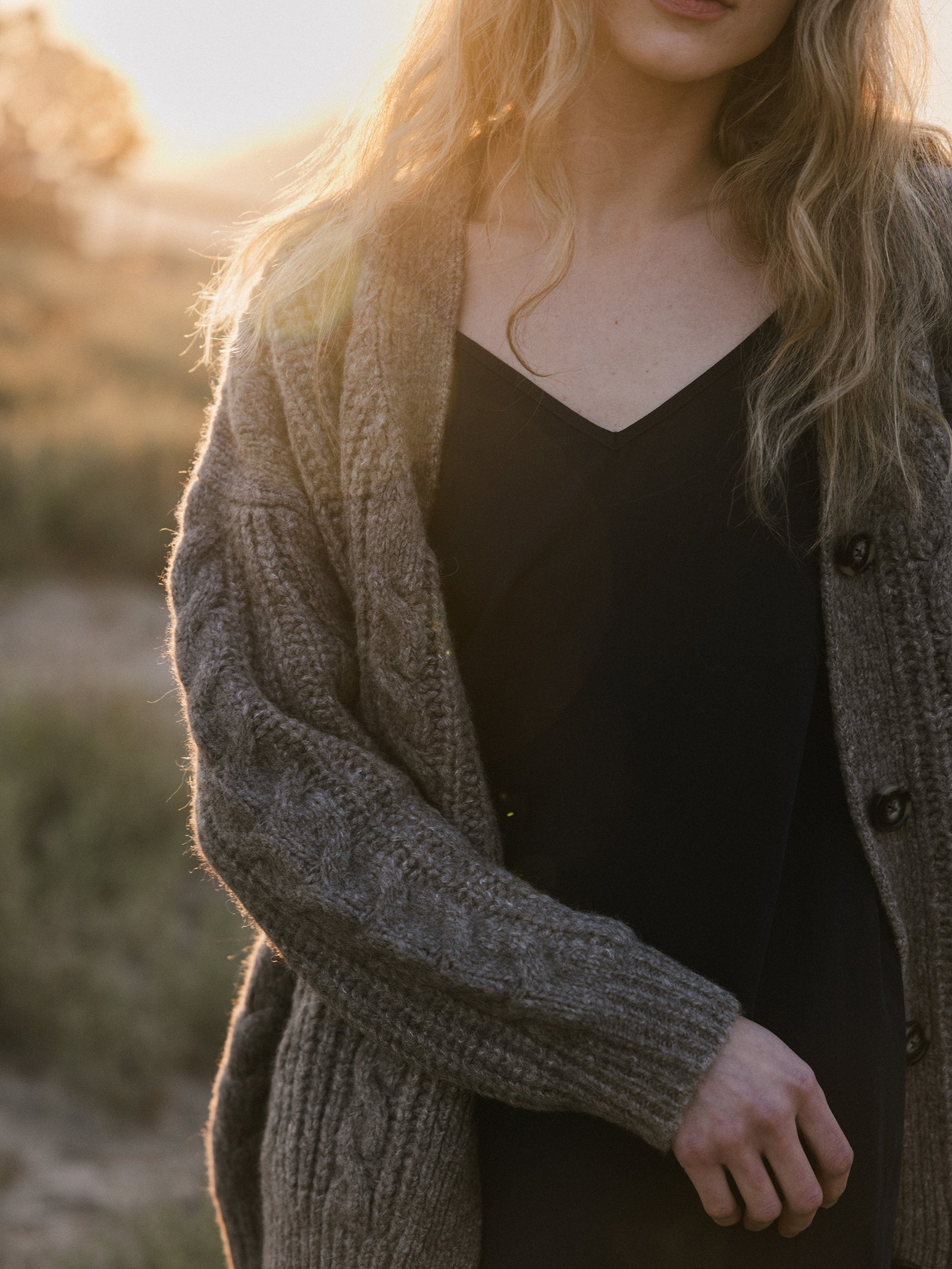 A woman with long blonde hair stands outdoors during golden hour, wearing a black dress and an Oversized Cable Knit Cardigan from Cozy Earth. The warm sunlight creates a soft glow, highlighting the natural setting with grass in the background. 