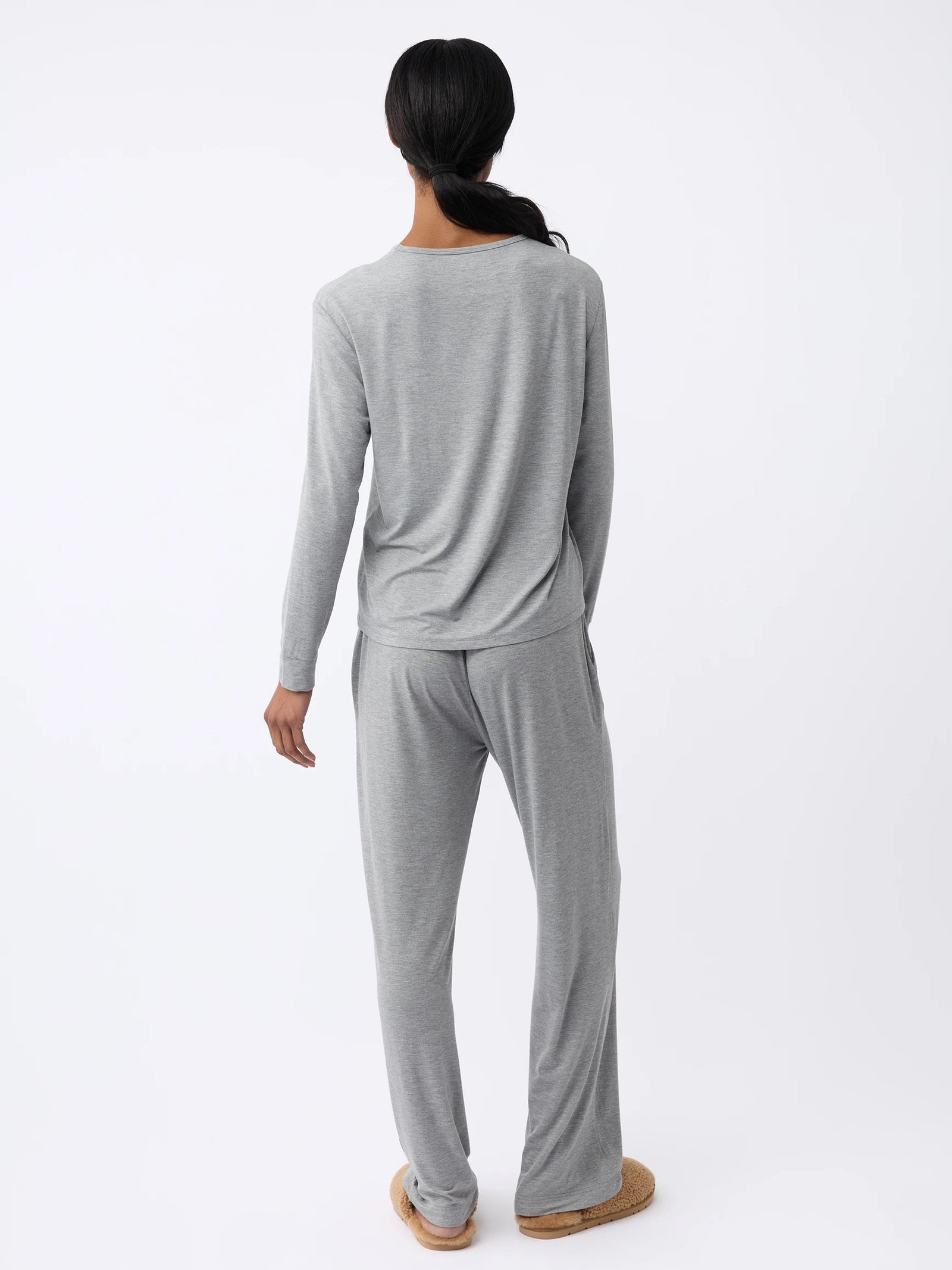 Someone with long hair, viewed from behind, wears a Cozy Earth Women's Stretch Knit Bamboo Long Sleeve Lounge Tee in gray with matching pants and slippers, standing against a light background. 