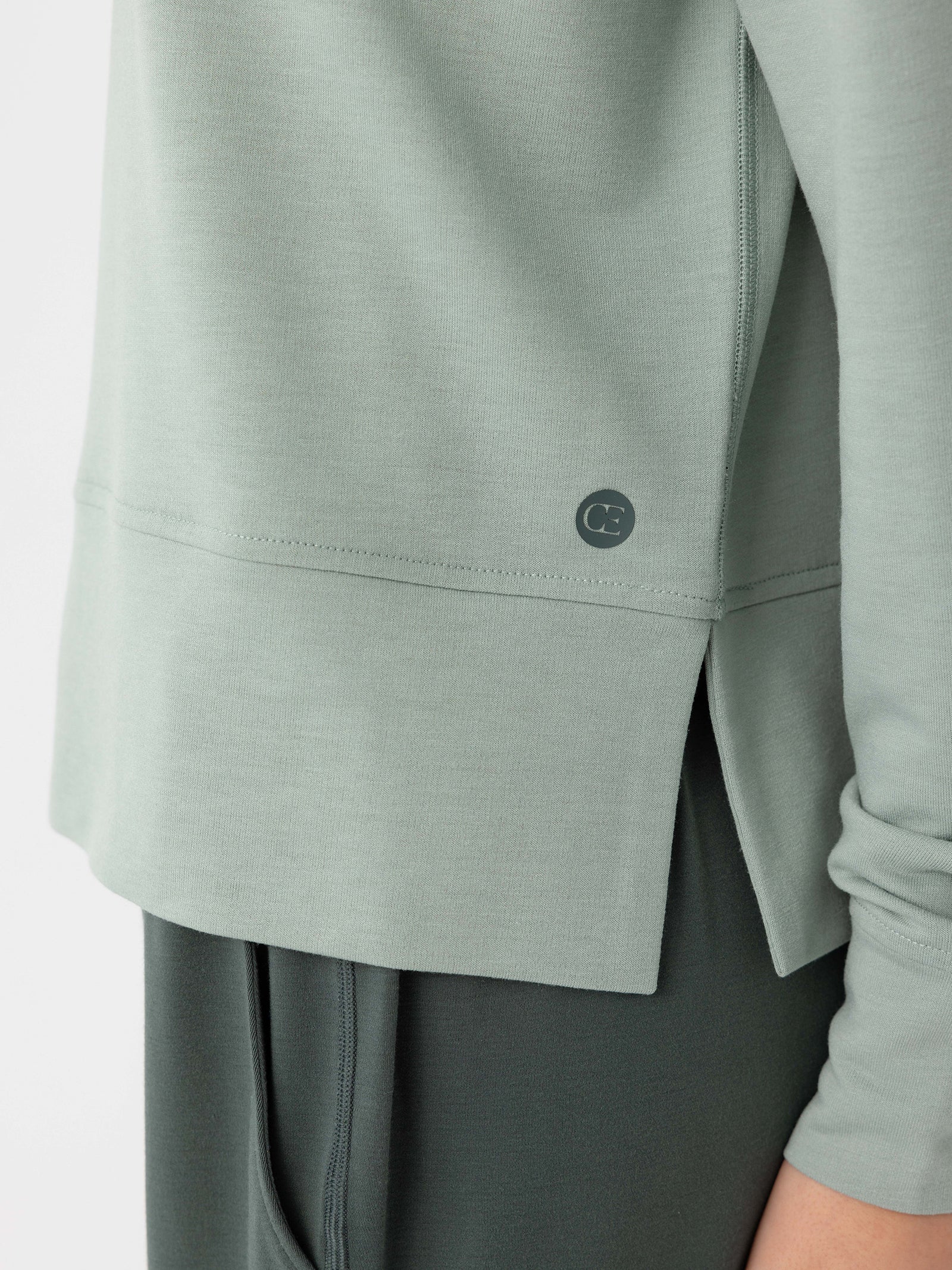 A close-up of a person wearing the Women's StudioLite Crewneck by Cozy Earth in light green, featuring a small circular logo near the hem on the left side. The shirt overlaps a pair of dark green pants, with an emphasis on the fabric texture and the side slit design. 