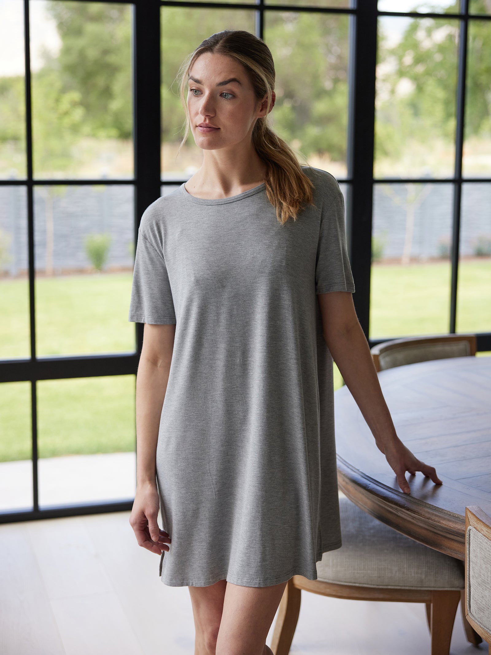 A woman in a Cozy Earth Women's Bamboo Stretch Knit Sleep Dress stands indoors by a wooden table. The gray, short-sleeve dress accentuates the natural light streaming through the large floor-to-ceiling windows, revealing a green lawn in the background. 