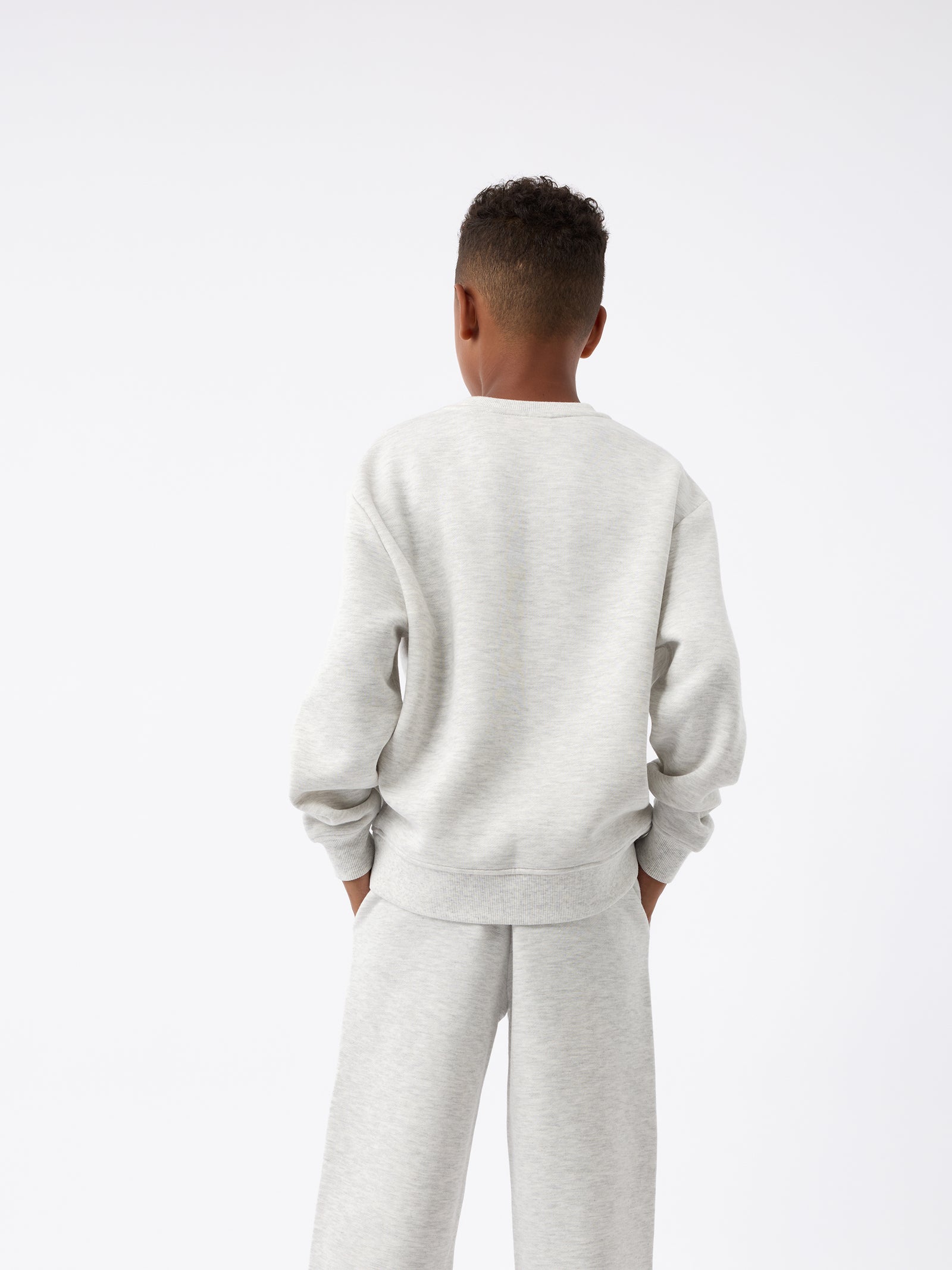 A child dressed in a light gray Kid's CityScape Crewneck from Cozy Earth, accompanied by matching pants, stands with their back to the camera. Their hands are tucked into their pockets against a plain white background. 